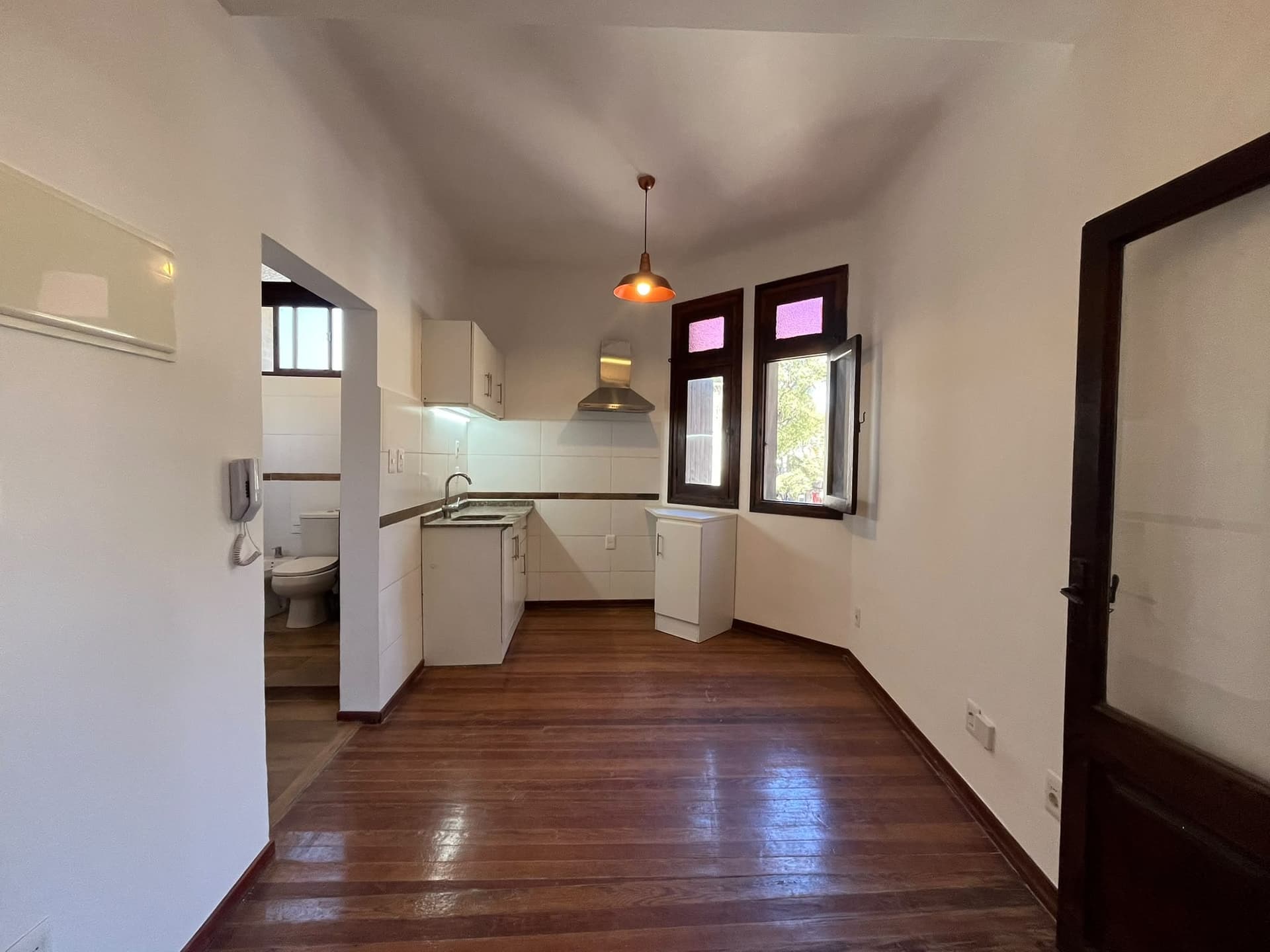 This image shows a combined kitchen and dining area with an adjacent bathroom visible through an open doorway. The kitchen features white cabinetry, a stainless steel sink, and a range hood, with white tiled backsplash. The room has dark wood flooring, white walls, and two dark-framed windows with an orange pendant light fixture.