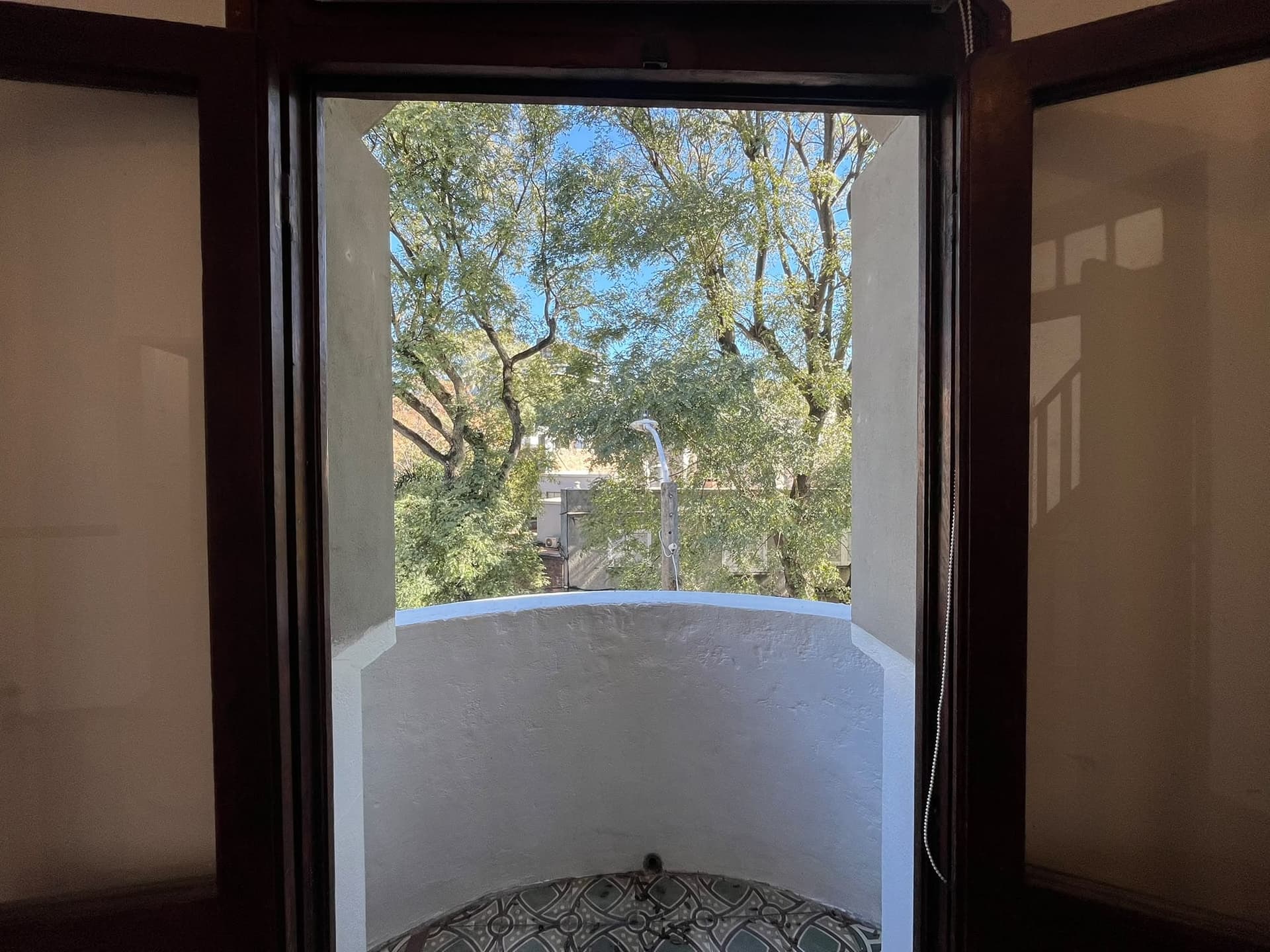 This image captures a view from an interior space looking out onto a small balcony or Juliet balcony. The dark wood frame of the opening contrasts with the light-colored stucco or painted wall of the balcony, which features decorative patterned tiles on its floor. Beyond the balcony, a vibrant view of lush green trees and a clear blue sky is visible, suggesting an outdoor urban or suburban setting.