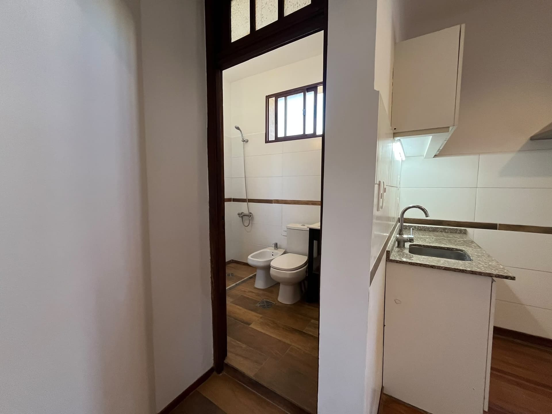 This image captures a partial view of a compact kitchen area with a granite countertop and stainless steel sink, adjacent to a doorway leading into a bathroom. The bathroom features white tiled walls, a toilet, a bidet, and a showerhead, with wood-look tile flooring visible in both spaces. A dark wood-framed window is present in the bathroom, and a dark wood door frame separates the two areas.