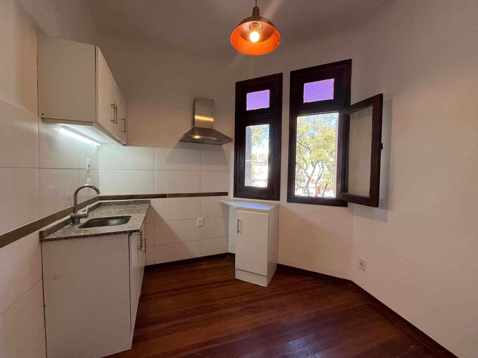 This is a compact kitchen featuring white cabinetry, a stainless steel range hood, and a granite countertop with an undermount sink. The room has dark wood flooring, white tiled walls, and two tall windows with dark frames, one of which is open to reveal an outdoor view. An orange pendant light fixture hangs from the ceiling.