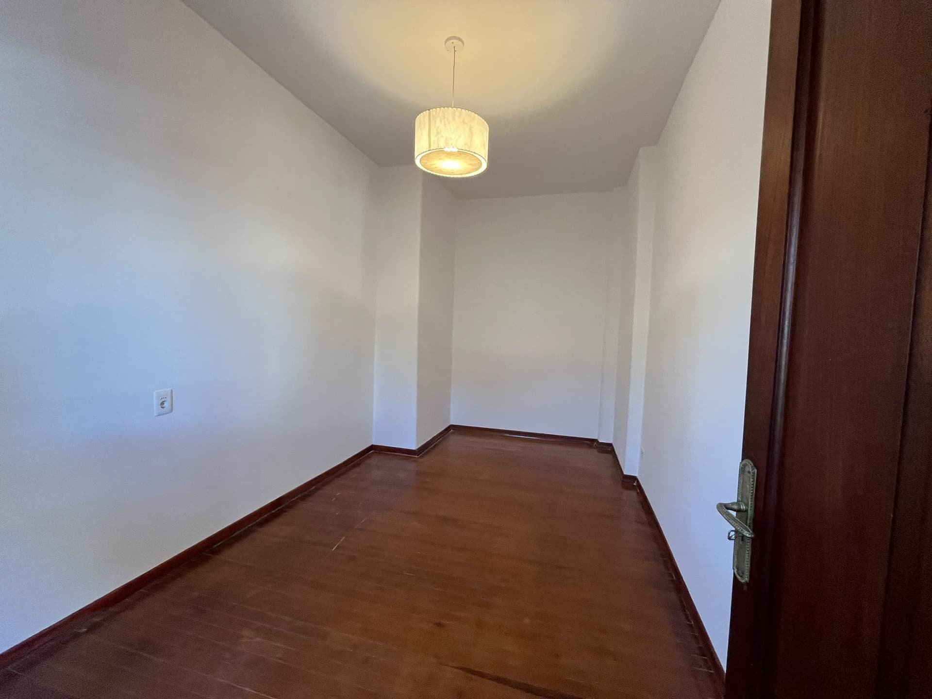 This is an empty room featuring white walls and dark hardwood flooring. A single light fixture with a drum shade hangs from the ceiling, providing ambient lighting. A dark wooden door with a vintage-style handle is visible on the right.