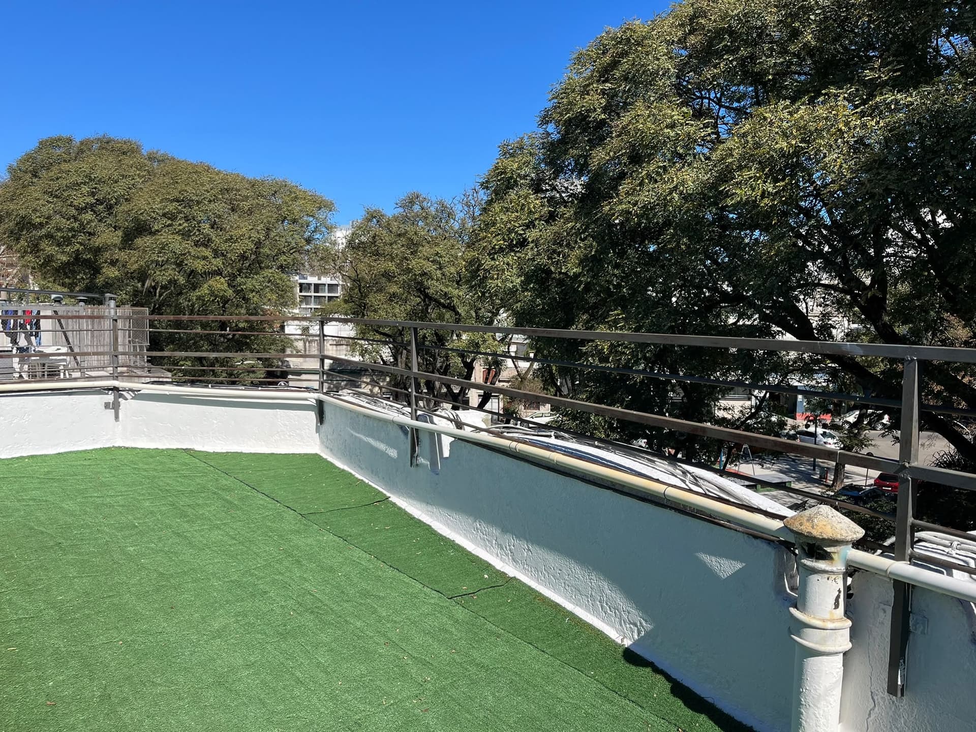 This image captures an outdoor rooftop space featuring artificial turf flooring and white painted parapet walls. A dark metal railing provides a boundary, overlooking a treelined street and distant buildings under a clear blue sky. A weathered pipe with a cap is visible in the foreground.