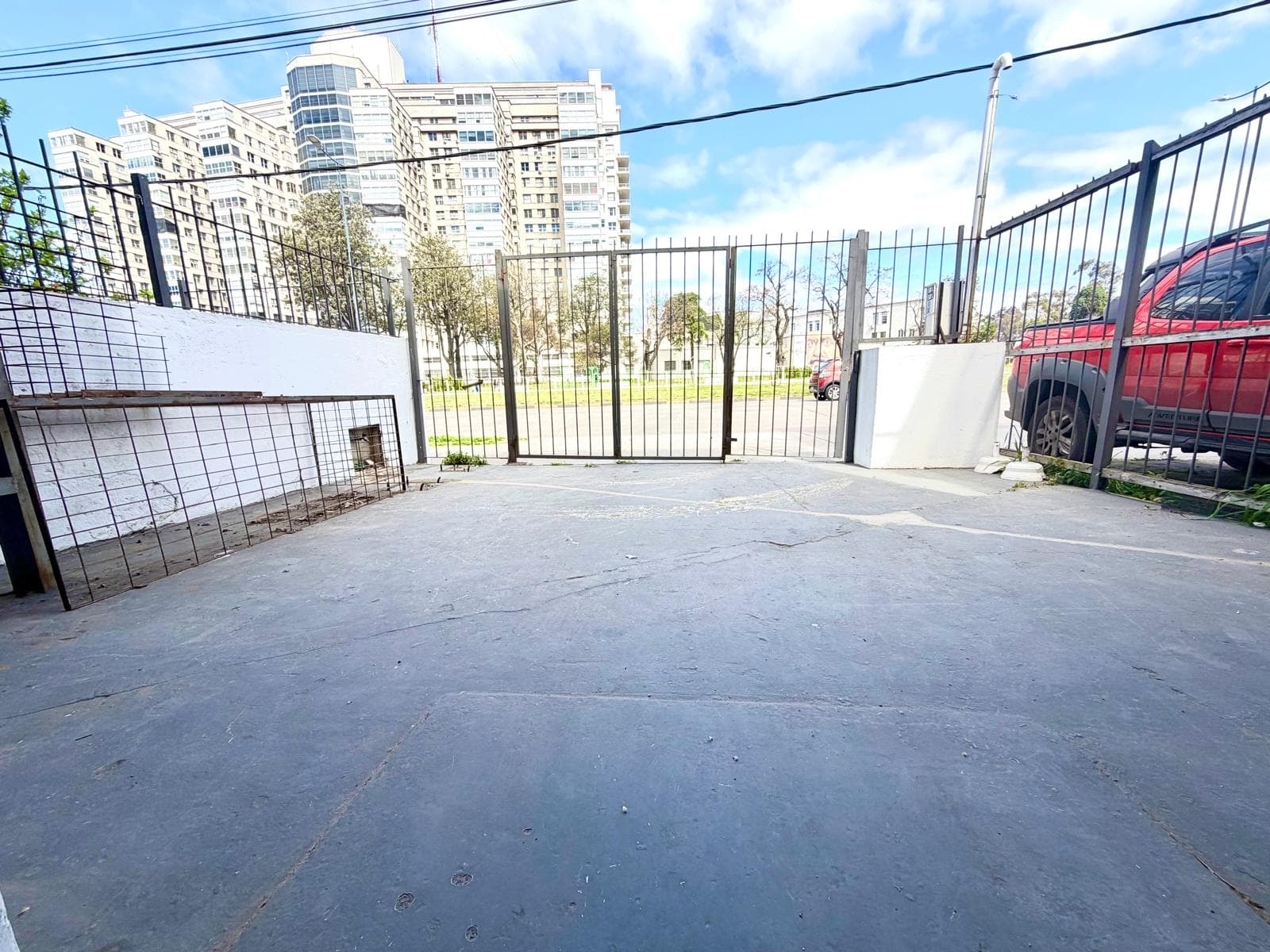 This exterior image shows a paved outdoor area, possibly a parking space or entrance, with a dark gray concrete surface. The area is enclosed by a black metal fence and gate, with a white wall on the left and a partial view of a red vehicle on the right. In the background, a large multi-story building and trees are visible under a partly cloudy sky.