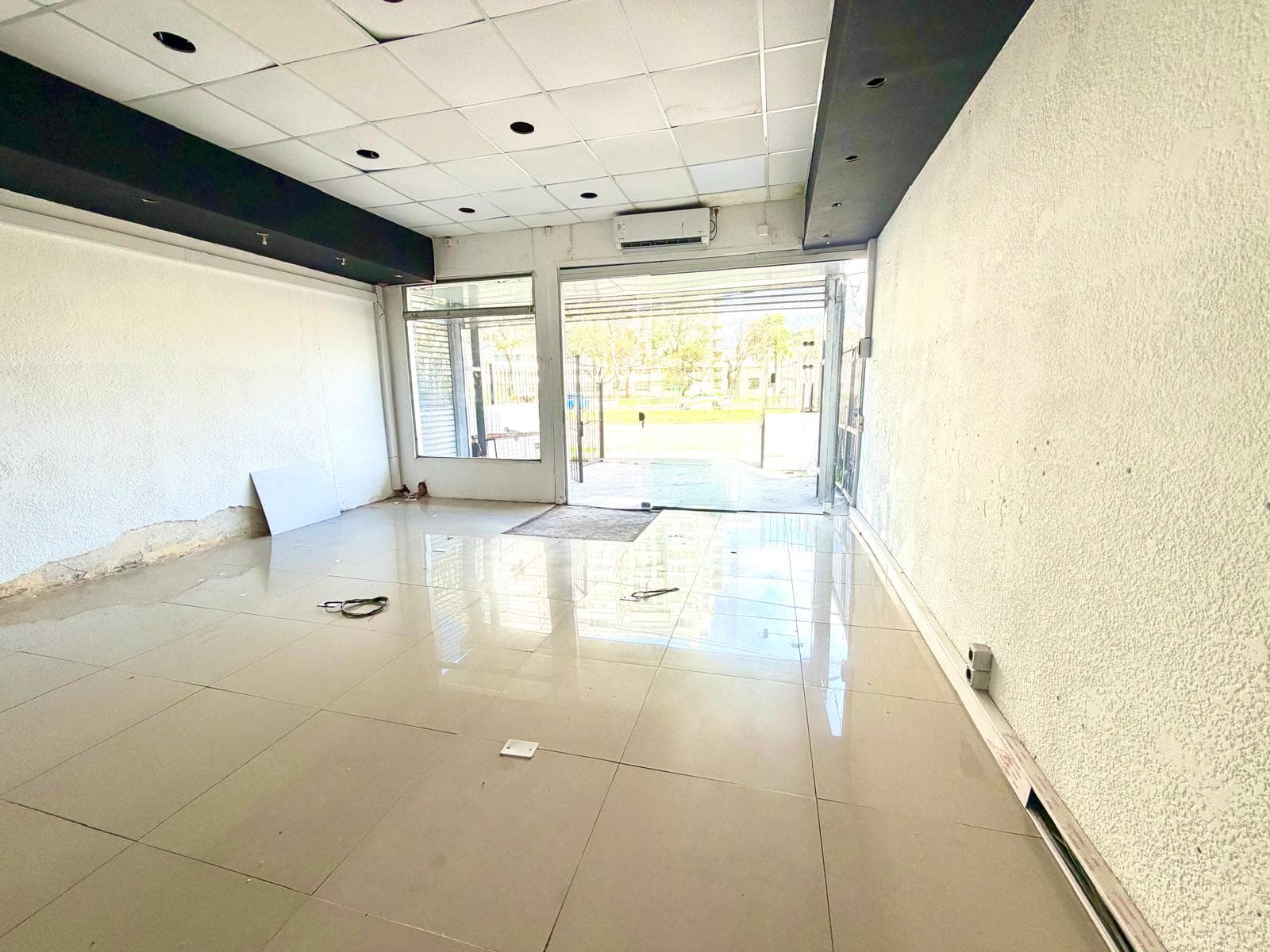 This is an interior view of a commercial space featuring light-colored tiled flooring and textured white walls. The space includes a dropped ceiling with recessed lighting and a visible air conditioning unit, with a large glass storefront providing natural light and a view of the exterior. The walls show some wear and tear, particularly near the base.