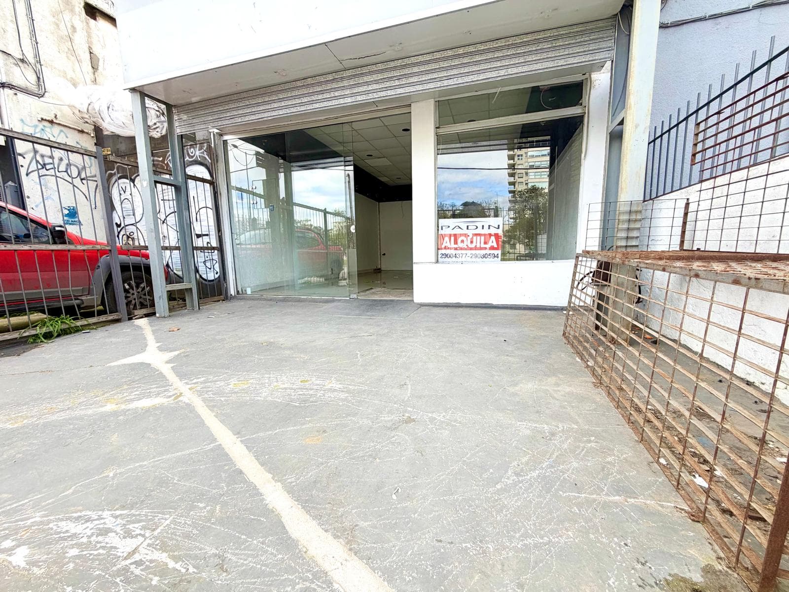 This is an exterior view of a commercial storefront available for rent. The space features large glass windows and a glass entry door, with a metal roll-down security shutter above. A "PADIN ALQUILA" sign is visible in the right window.
WATERMARK: PADIN