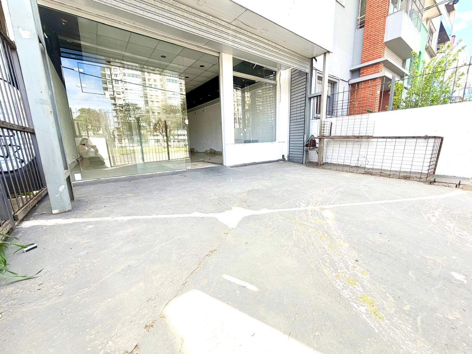 Exterior view of a commercial storefront with a large glass facade and an adjacent roll-down security shutter. The concrete ground in front shows signs of wear and some discoloration, with a metal grate leaning against the building on the right. The interior appears vacant and ready for customization.
