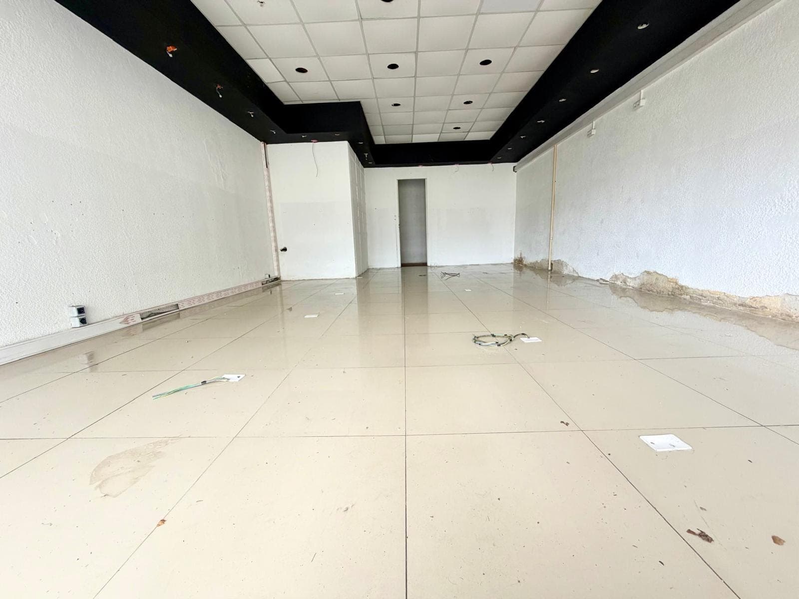 This is an interior view of a commercial space or large room, currently in a state of renovation or disrepair. Key features include light-colored tiled flooring, white textured walls with visible water damage and exposed wiring, and a drop ceiling with recessed lighting. A black bulkhead feature runs along the ceiling, and an open doorway is visible at the far end.