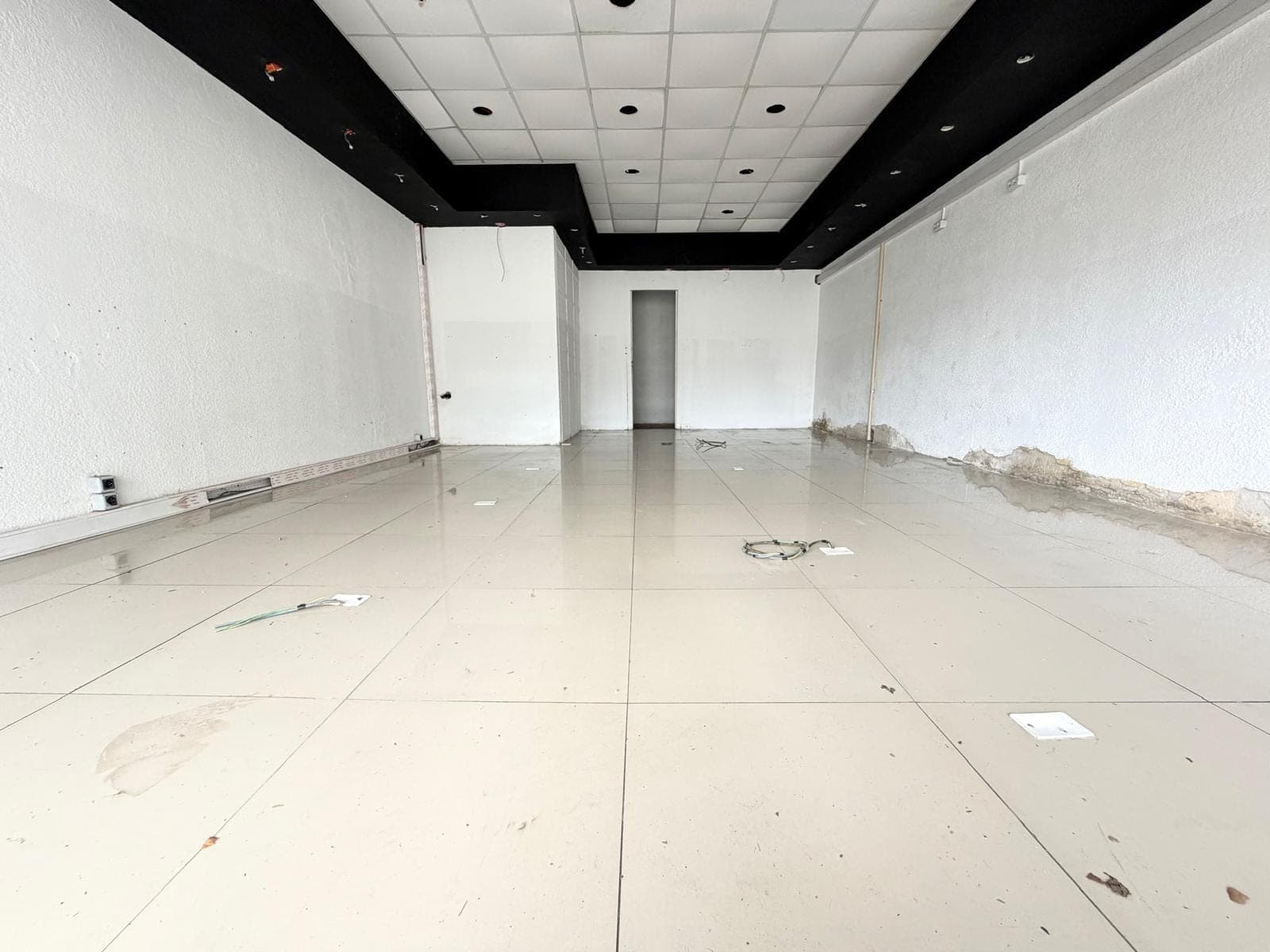 This is an interior view of a commercial space, currently in a state of disrepair or renovation. Key features include white textured walls, a drop ceiling with recessed lighting, and a black soffit detail. The light-colored tiled floor shows signs of wear and debris, and there is visible damage to the lower portion of the walls.