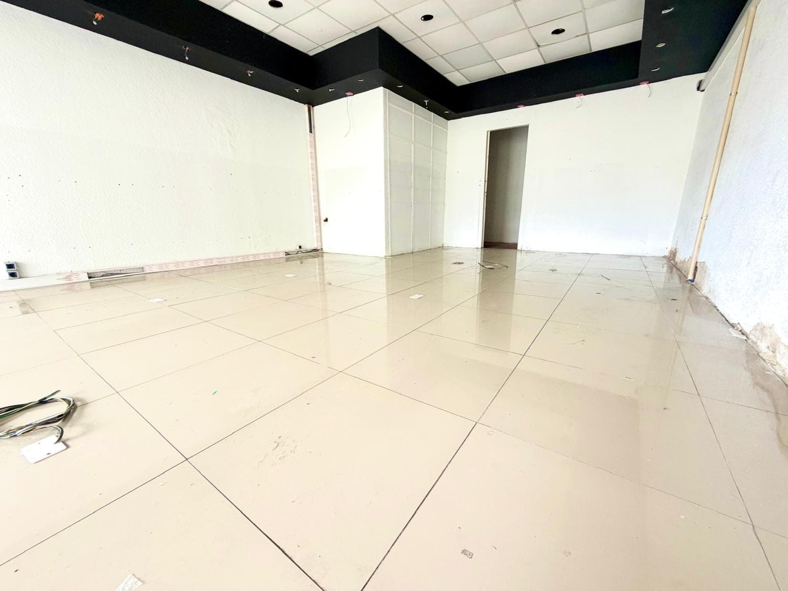 This is an interior view of a commercial space or large room, currently unfurnished and undergoing renovation or preparation. Key features include light-colored tiled flooring, white walls, and a black ceiling soffit with recessed lighting, contrasting with a white grid ceiling. Exposed wiring and unfinished wall sections indicate ongoing work.