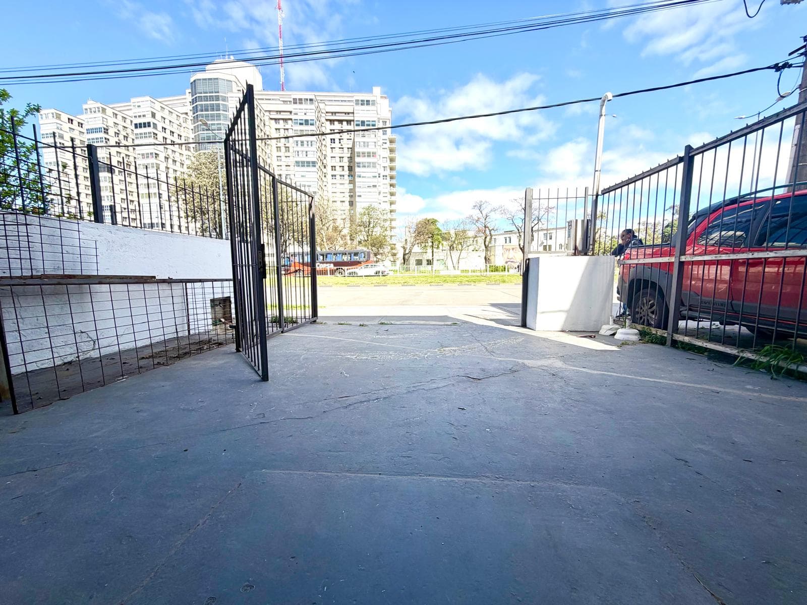 This image captures an exterior view of a paved entrance area, likely a driveway or parking space, with an open black metal gate. The concrete ground shows some wear and cracks, and a white wall with a metal grid fence runs along one side. In the background, a large white multi-story building and a red pickup truck are visible under a bright blue sky with scattered clouds.