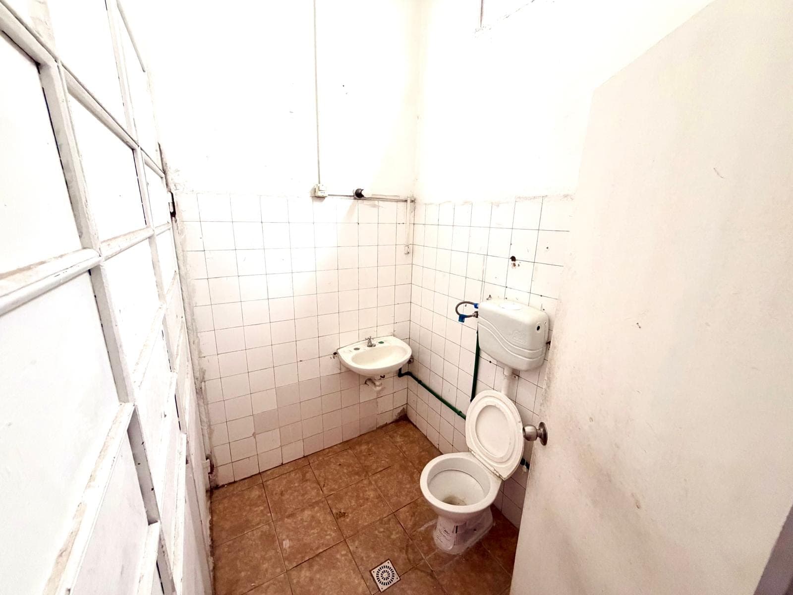 This is a compact bathroom featuring a white toilet with an exposed cistern and a wall-mounted sink. The walls are partially tiled with white ceramic tiles, and the floor is covered with brown tiled flooring. The room appears to be in fair condition, showing some signs of wear and exposed plumbing.