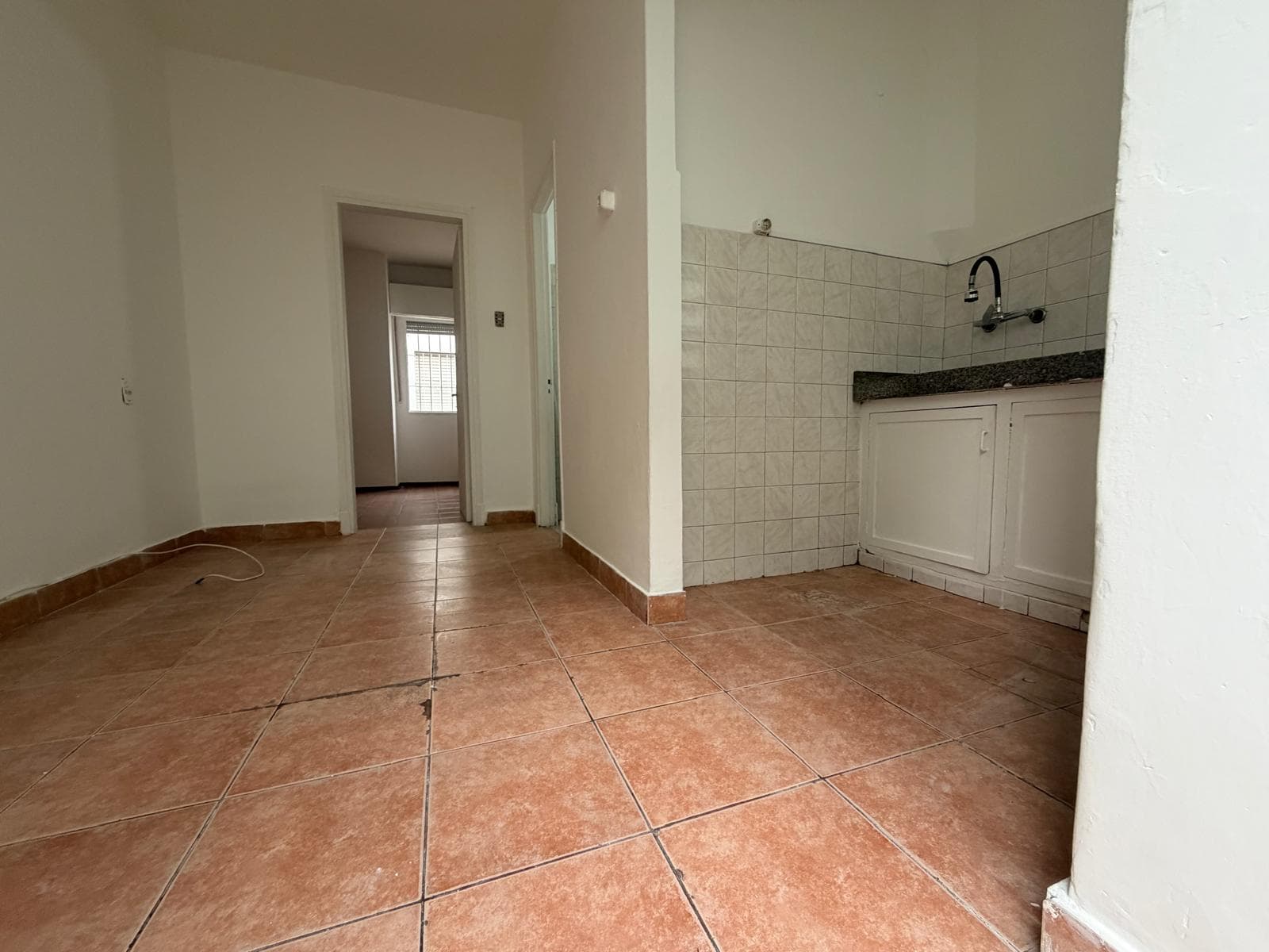 This image shows a compact kitchen area within a larger room, featuring terracotta-colored floor tiles and white painted walls. The kitchen section includes a tiled backsplash, a dark granite-look countertop, a sink with a black flexible faucet, and white base cabinets. A doorway leading to another room with a window is visible in the background.