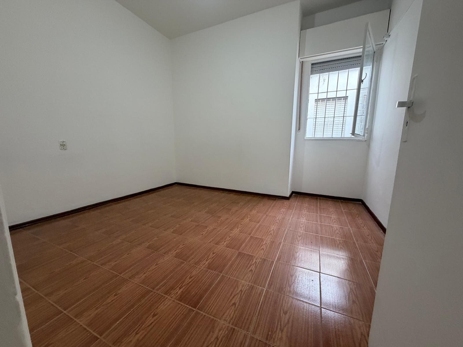 This is an empty bedroom featuring light-colored walls and brown tiled flooring with dark baseboards. A window with security bars and an open casement provides natural light. The room appears clean and ready for occupancy.