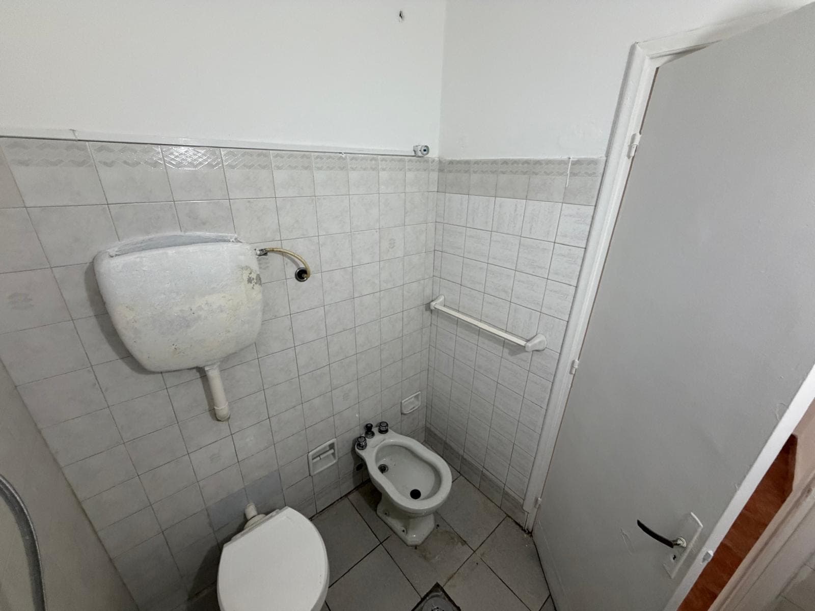 This is a small, tiled bathroom featuring a toilet with an exposed, high-mounted cistern, and a bidet. The white tiled walls show some wear and discoloration, particularly on the cistern, and there is a white grab bar installed above the bidet. A white door with a black handle is visible on the right.