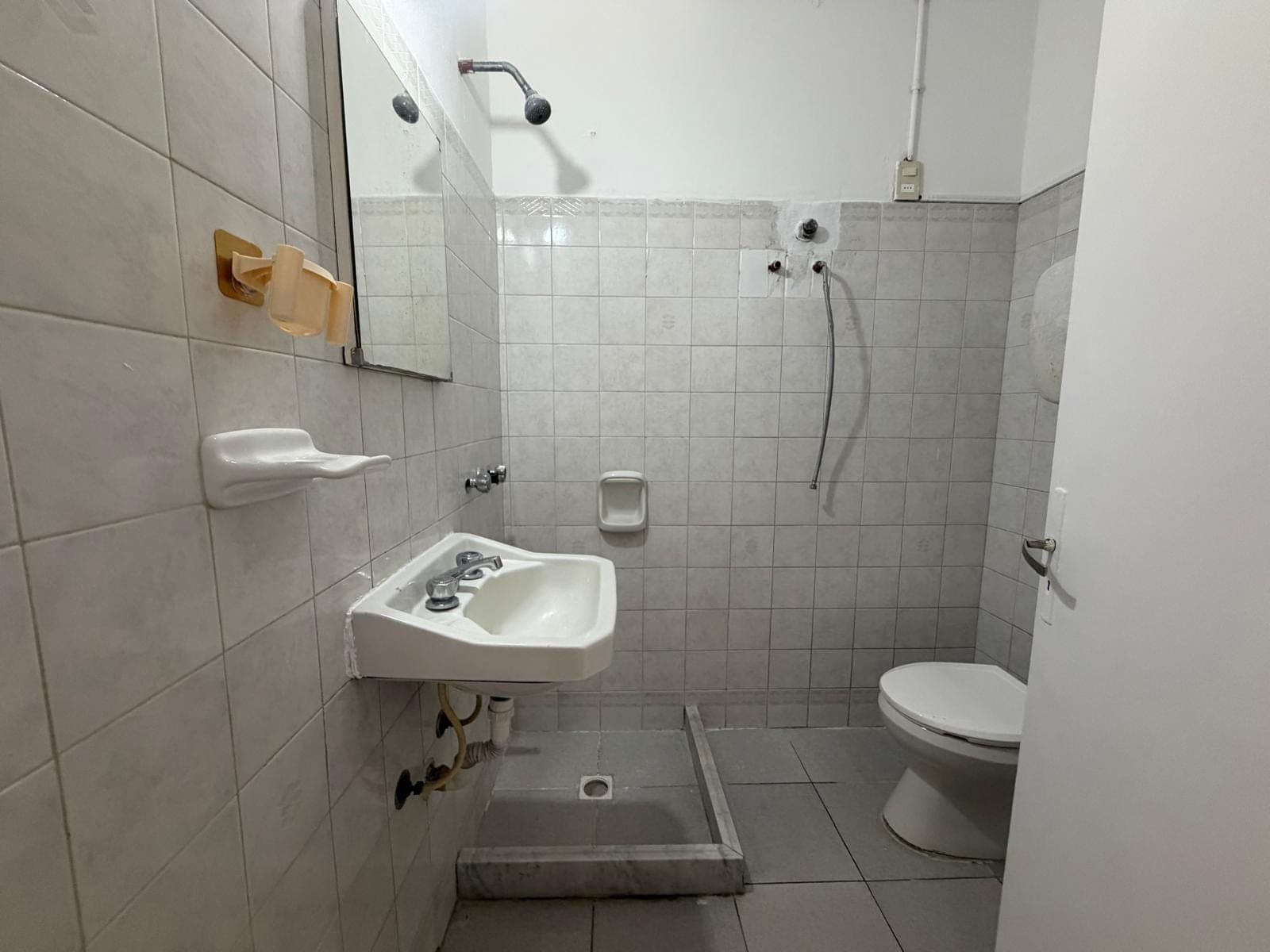 This is a functional bathroom featuring light-colored tiled walls and floor. Key features include a wall-mounted sink with exposed plumbing, a toilet, and a showerhead. The room appears to be in fair condition, with some visible wear and tear on the tiles and fixtures.