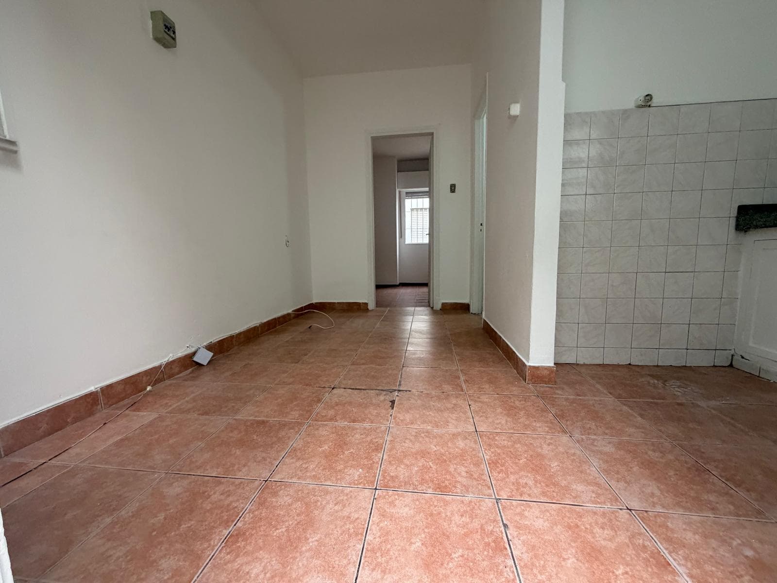 This image depicts an empty room, possibly a living area or entry space, featuring terracotta-colored floor tiles and white painted walls. A partial tiled wall, likely indicating a kitchen or utility area, is visible on the right, while a doorway leads to another room with a window at the far end. The space appears to be in fair condition, ready for renovation or furnishing.
