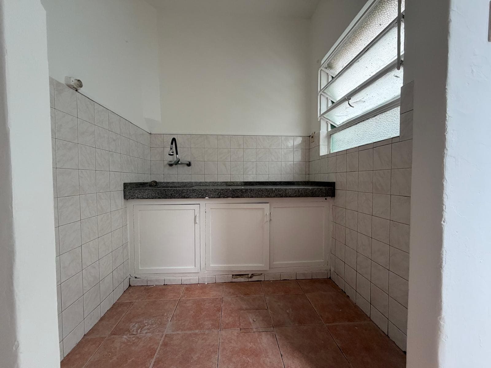 This is a utility room or small kitchen space featuring white tiled walls and a dark granite-look countertop with an integrated sink and faucet. White base cabinets are installed below the counter, and the room has terracotta-colored floor tiles. A louvered window with textured glass provides natural light.