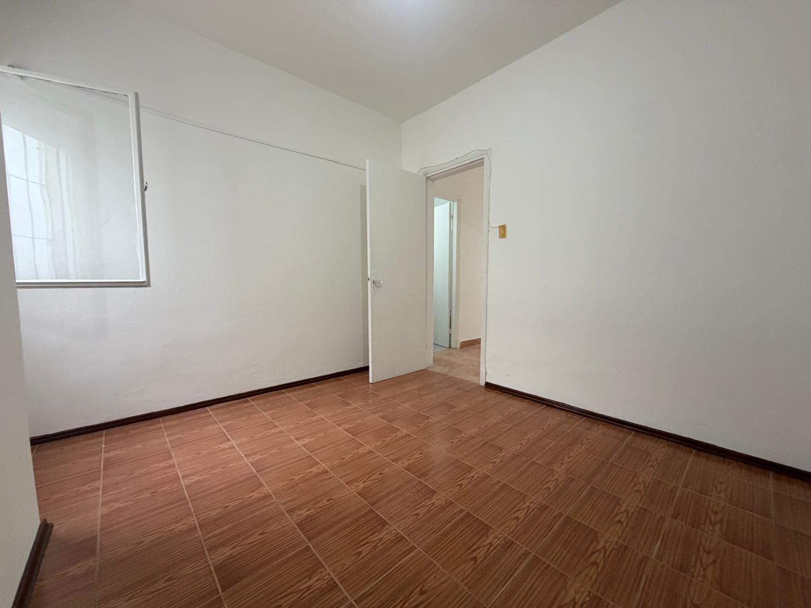 This is an empty room, likely a bedroom or office, featuring white painted walls and brown tiled flooring. A window with a white frame is visible on the left wall, and an open white door leads to another room with similar flooring. The room appears clean and well-maintained.