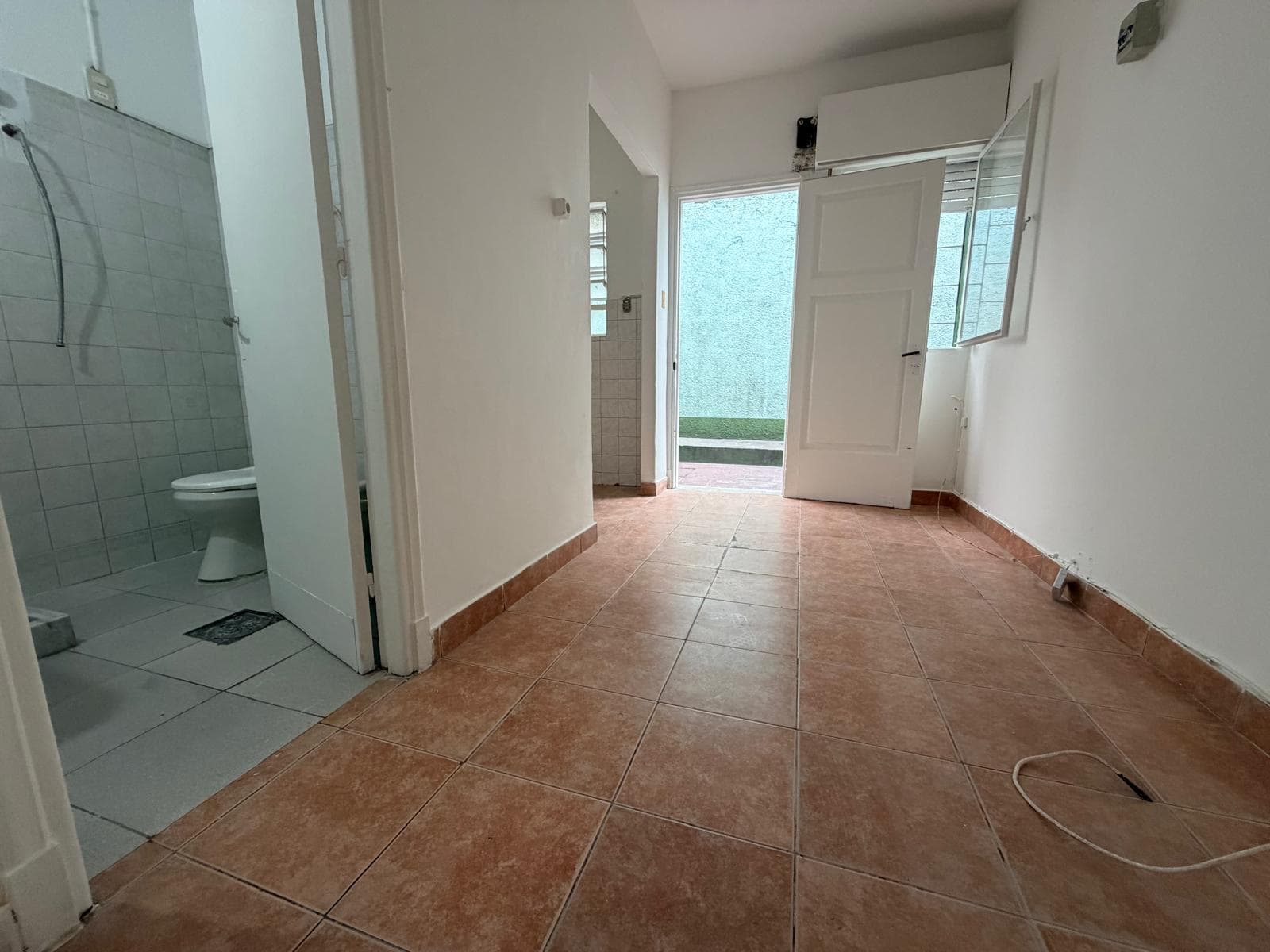 This image shows a multi-purpose room with terracotta tile flooring and freshly painted white walls. To the left, a partial view of a bathroom with light gray tiled walls and a toilet is visible. An open door leads to an exterior space with green artificial turf.