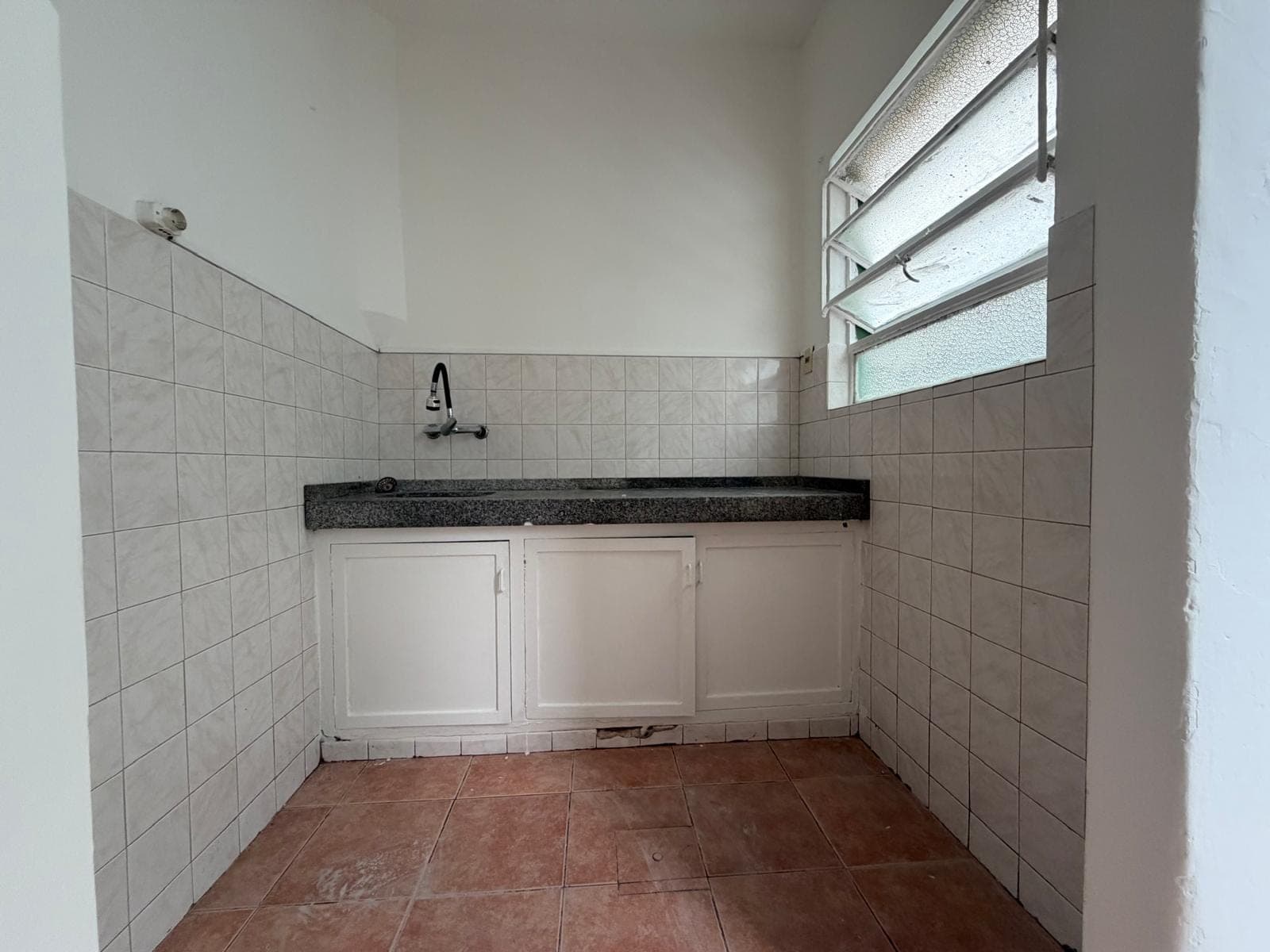 This is a compact kitchen space featuring white tiled walls, a dark granite countertop with an integrated sink and faucet, and white base cabinets. The room has terracotta-colored floor tiles and a louvered window providing natural light. The overall condition appears functional but dated.
