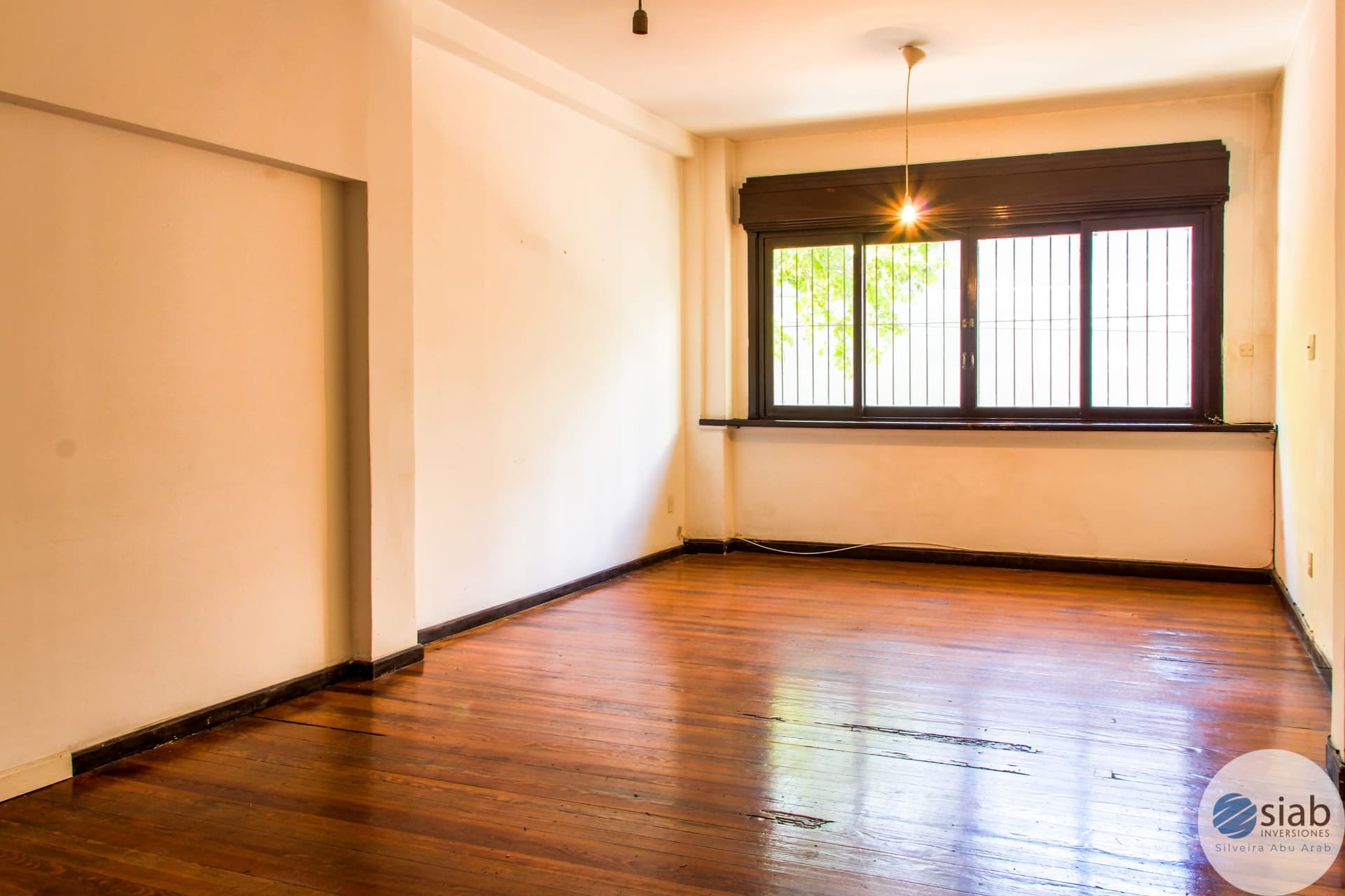 # Property Description
This is a spacious living room or bedroom featuring warm cream-colored walls and hardwood flooring in excellent condition. The room is flooded with natural light from a large multi-pane window with dark wood framing that overlooks greenery, and is complemented by pendant lighting fixtures. The space offers clean lines, neutral décor, and ample room for furniture arrangement, making it ideal for residential use.
WATERMARK: SIAB Inversiones - Silveira Abu Arab