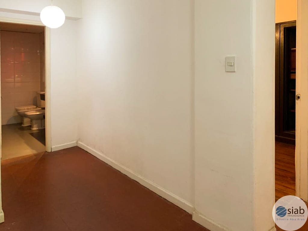 This is a hallway or corridor space featuring clean cream-colored walls, dark reddish-brown flooring, and bright overhead lighting from a modern pendant fixture. The space provides access to multiple rooms, including a bathroom visible on the left and additional doorways on the right, with warm wood-toned trim throughout. The area is well-maintained and appears to be part of a residential apartment or unit.
WATERMARK: SIAB Investimentos