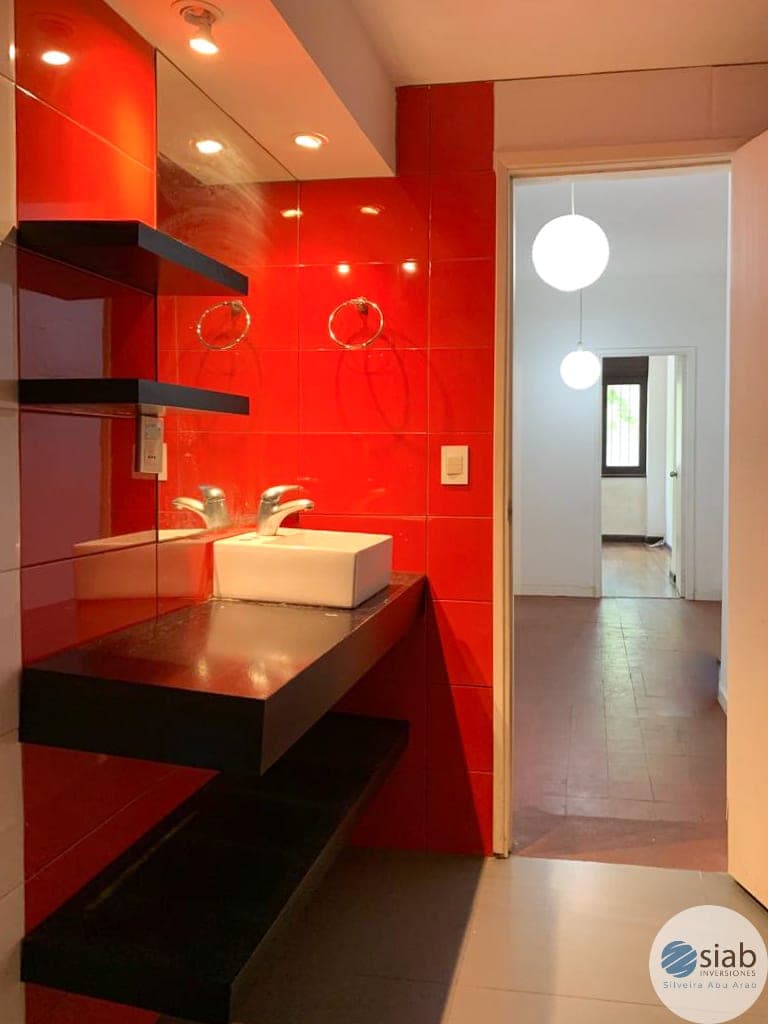 # Bathroom Description
Modern bathroom featuring a striking red tile accent wall with contemporary design elements. The space includes a floating black vanity with a white vessel sink, brass faucet fixtures, and integrated recessed lighting throughout the ceiling. Pendant globe lights hang above the vanity, and the room opens to an adjacent hallway with brown tile flooring, creating a bright, well-lit space with bold aesthetic appeal.
WATERMARK: SIAB Imobiliário