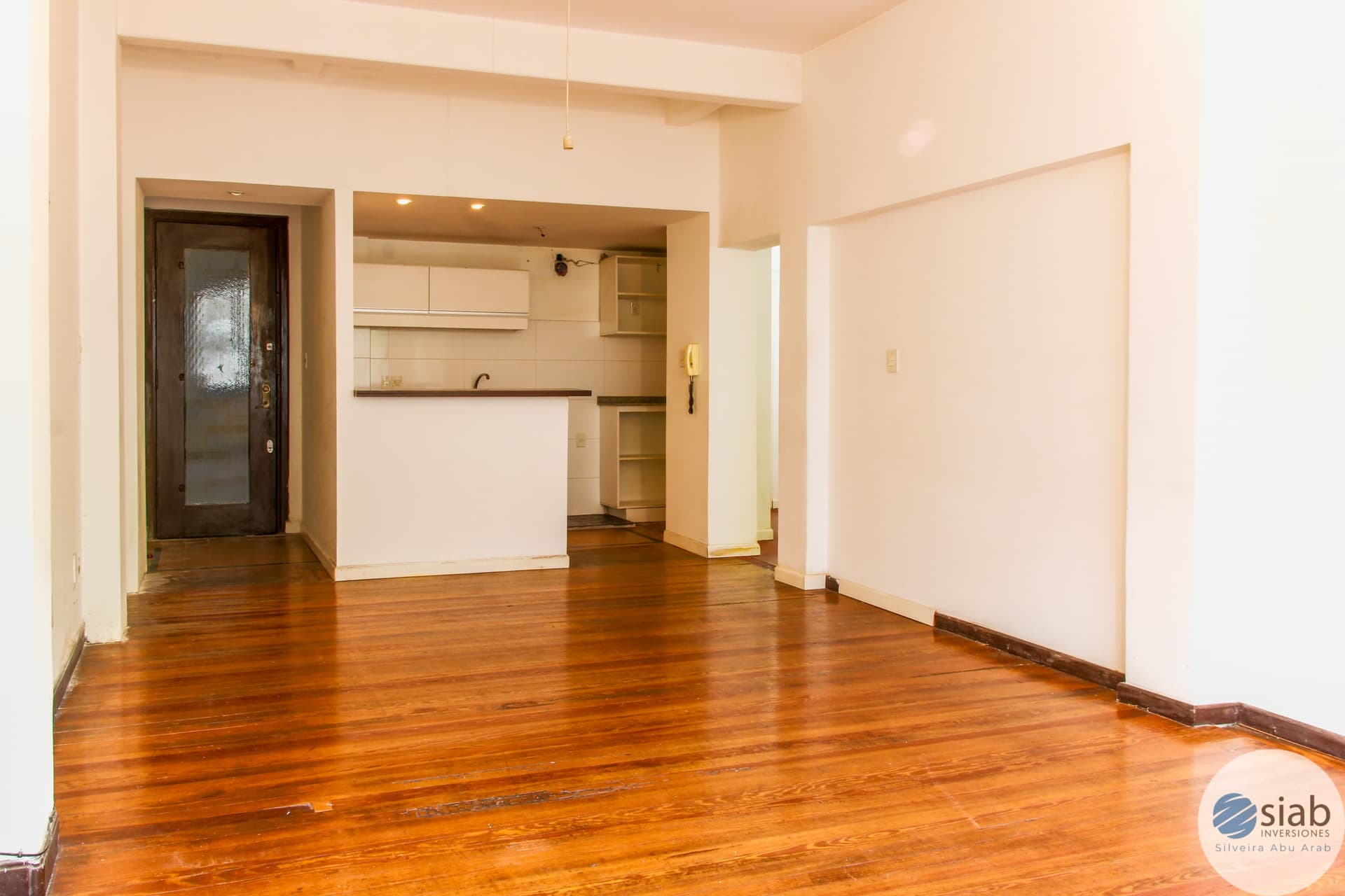 # Property Description
This is a spacious open-concept living and kitchen area featuring warm hardwood flooring throughout. The kitchen includes white cabinetry, built-in shelving, recessed lighting, and a breakfast counter, while the adjoining living space offers ample room and natural light. The unit includes an entry foyer with a dark wood door and provides excellent flow between functional areas.
WATERMARK: SIAB Inversiones - Silveira Abu Arab