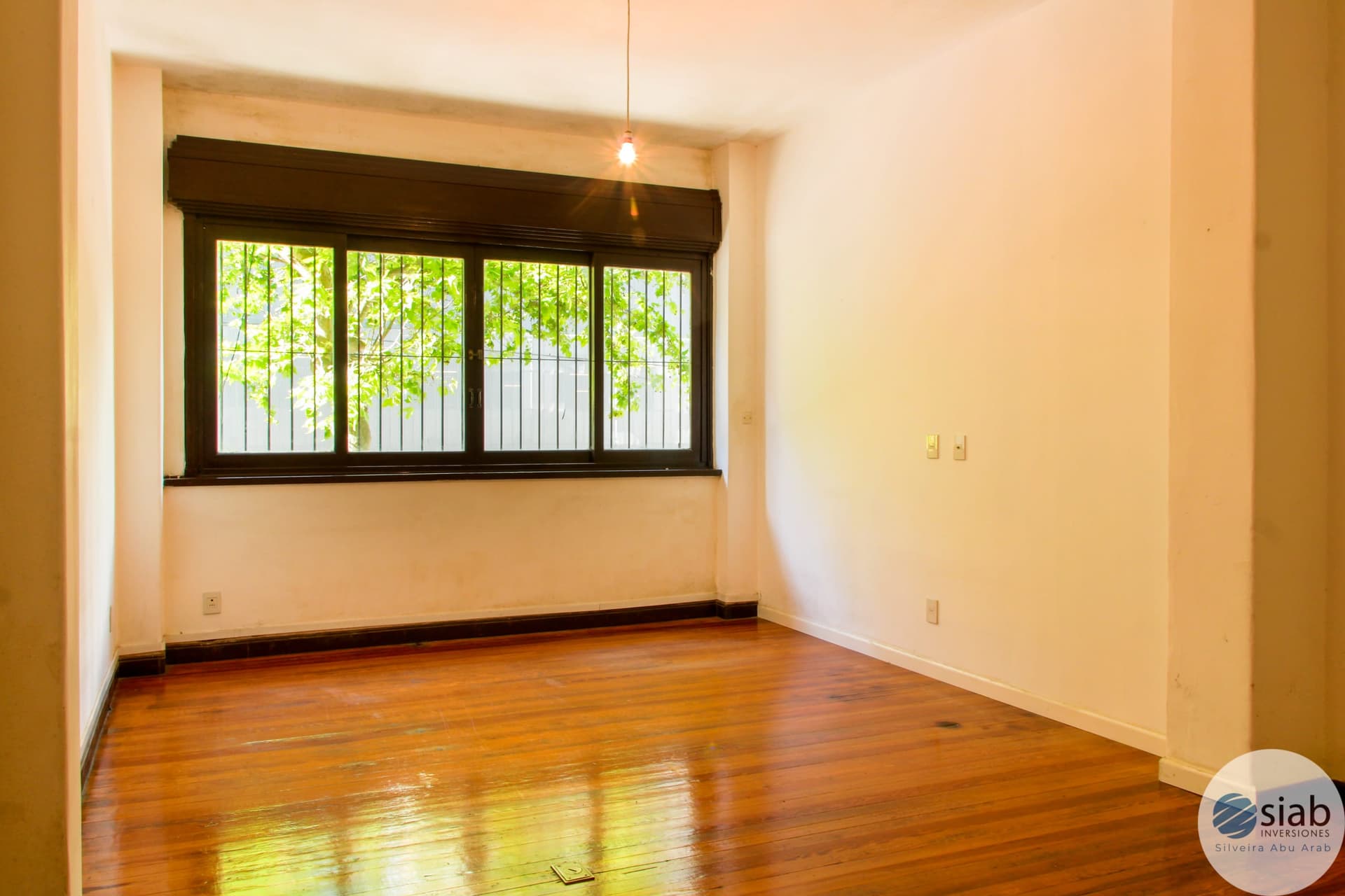# Property Description
This is a bright, spacious living room or bedroom featuring warm cream-colored walls and beautiful hardwood flooring in excellent condition. The room is flooded with natural light from a large four-panel dark-framed window with security bars, offering views of lush green foliage and a white fence. The space includes a pendant light fixture, electrical outlets, and clean, neutral finishes that create an airy, well-maintained interior.
WATERMARK: SIAB Inversiones - Silveira Abu Arab