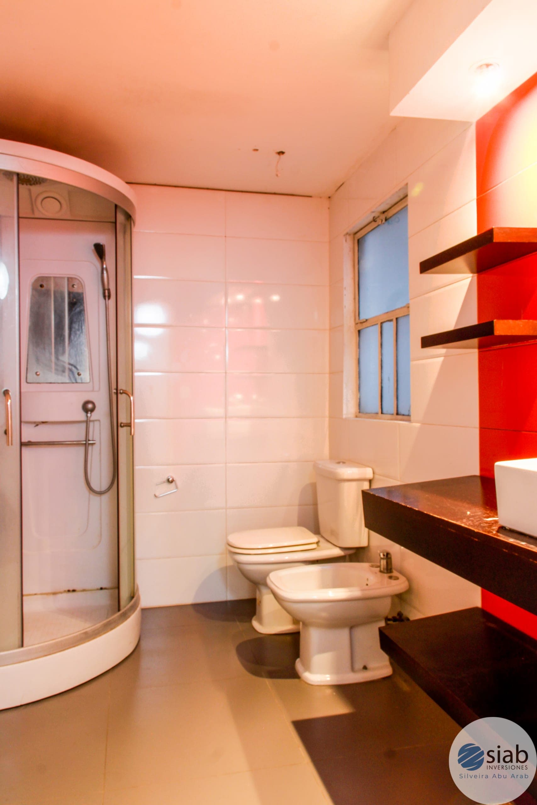 # Bathroom Description
Modern bathroom featuring a curved glass shower enclosure with chrome fixtures on the left side, complemented by cream-colored ceramic tile walls and a polished concrete floor. The space includes a toilet, bidet, and dark wood vanity with red accent shelving on the right wall, along with a small window providing natural light. The bathroom is well-appointed with contemporary design elements and appears to be in excellent condition.
WATERMARK: SIAB Inversiones - Silveira Abu Arab