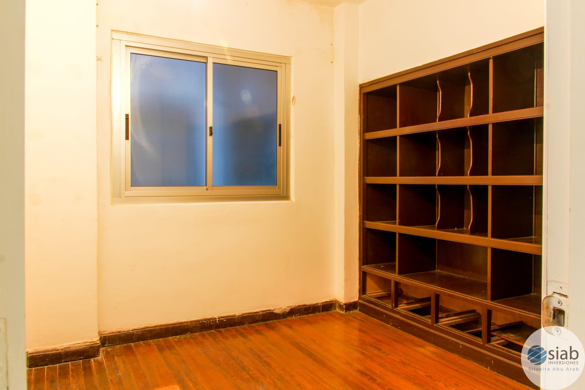 # Property Image Description
This is a bedroom or study space featuring warm cream-colored walls and a polished wooden floor in a rich amber tone. The room includes a large wooden sliding window with natural light exposure and built-in wooden shelving units with multiple compartments along one wall, providing substantial storage. The space appears clean and well-maintained with simple, functional design elements.
WATERMARK: SIAB Inversiones (Silveira Abu Arab)