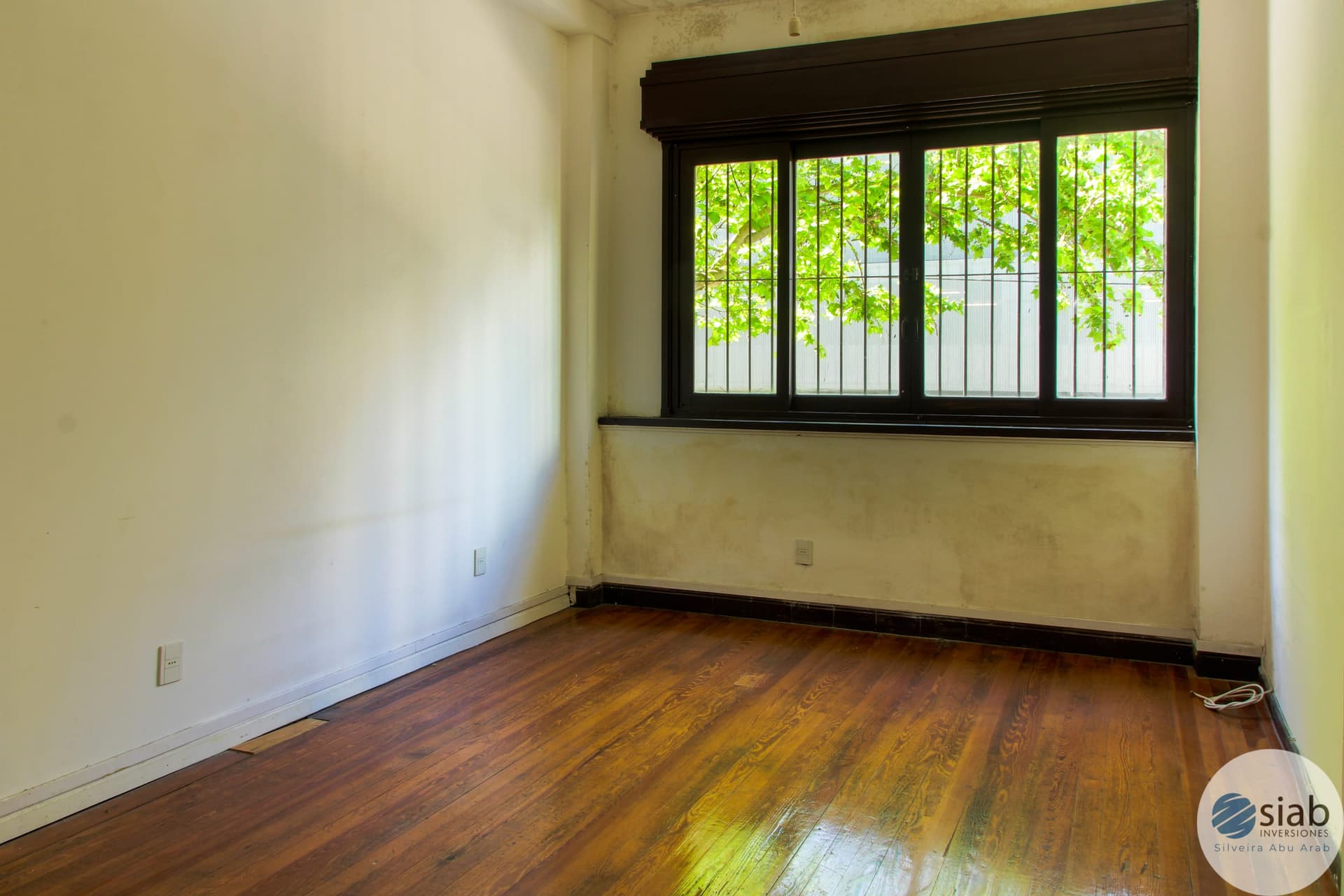 # Property Image Description
This is a bright, spacious bedroom or living room featuring hardwood flooring in warm medium-to-dark tones and cream-colored walls. The room is flooded with natural light from a large four-panel window with dark wood framing that overlooks lush green foliage and a security gate, creating a serene garden view. The space includes electrical outlets and appears to be in good condition, offering excellent natural ventilation and a peaceful ambiance.
WATERMARK: SIAB Inversiones (Silveira Abu Arab)