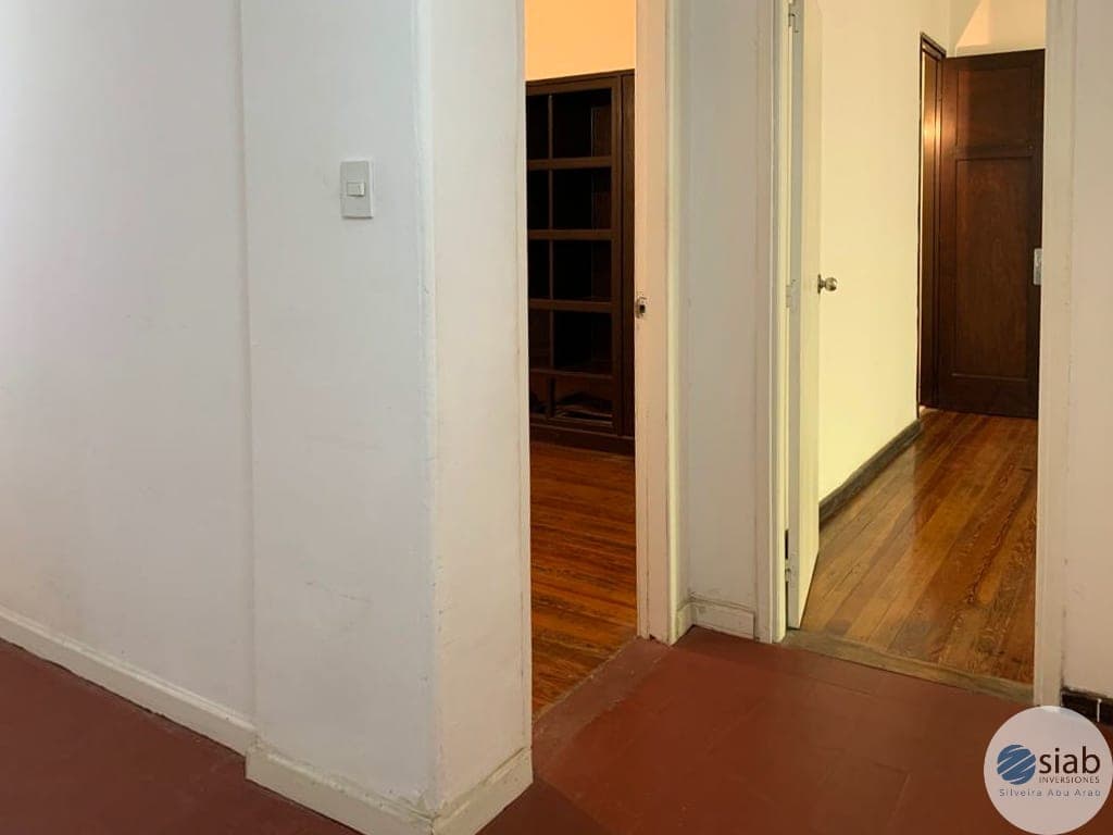 This image shows a hallway or corridor area with mixed flooring, featuring a red/terracotta tile section in the foreground transitioning to warm-toned wood flooring. The space includes white painted walls, multiple doorways with wooden doors, and built-in wooden shelving visible through one of the open doors. The hallway is well-lit with warm lighting and appears to be in good condition with clean, maintained surfaces.
WATERMARK: SIAB Inversiones