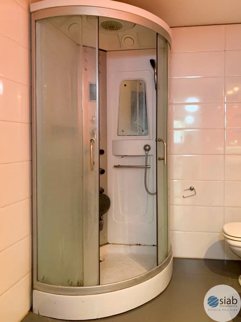 # Bathroom - Shower Enclosure
This bathroom features a modern curved corner shower enclosure with frosted glass sliding doors and an overhead rainfall showerhead. The space includes light pink tile walls, a white toilet, and chrome fixtures in good condition. The shower unit is well-maintained with a compact, space-efficient design suitable for smaller bathrooms.
WATERMARK: SIAB Imobiliárias