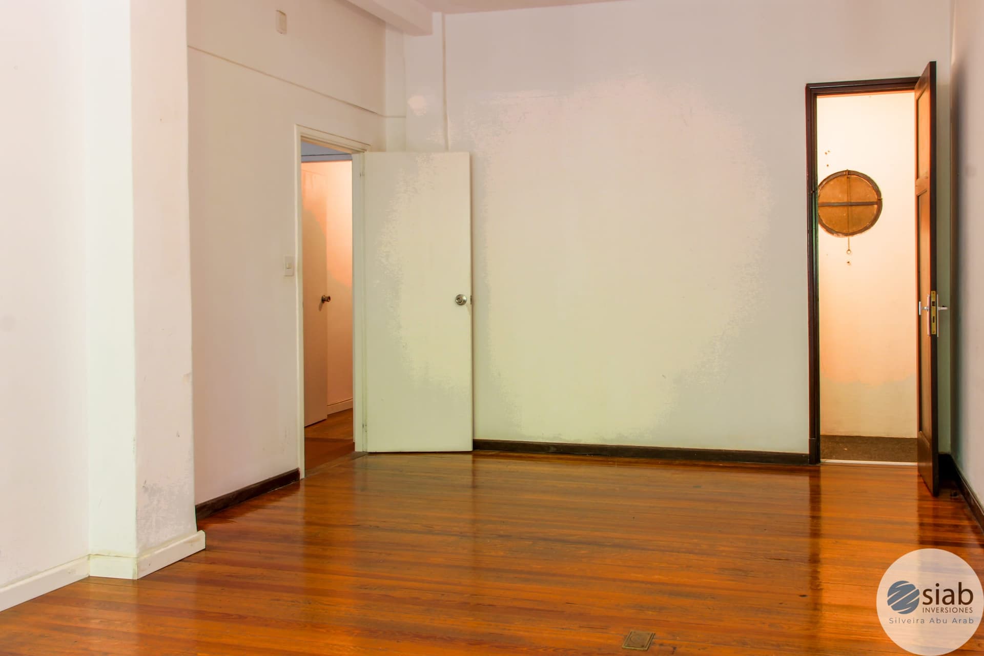 # Property Description
This is a spacious interior room with warm hardwood flooring and clean white walls, featuring multiple doorways providing access to adjacent spaces. The room includes built-in closet storage with light-colored doors and abundant natural light from glass-paned doors on the right side. The polished wood floors and neutral finishes create a bright, airy atmosphere suitable for various uses such as a bedroom, living area, or multi-purpose space.
WATERMARK: SIAB Inversiones - Silveira Abu Arab