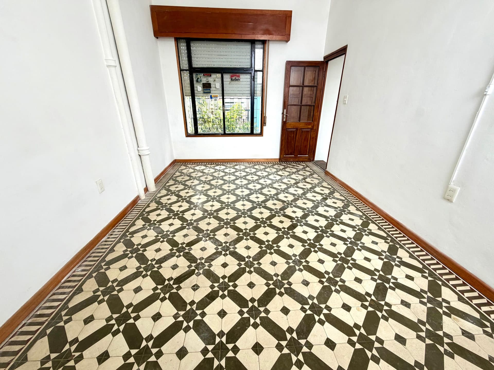 This is a spacious interior room or hallway featuring distinctive black and cream geometric patterned tile flooring with an ornamental border. The space has clean white walls, a wooden-framed window with security bars offering natural light, and a solid wooden door with six panels. The room is accented with warm wood trim throughout and includes basic electrical outlets, presenting a well-maintained, traditional aesthetic suitable for a residential or commercial property.