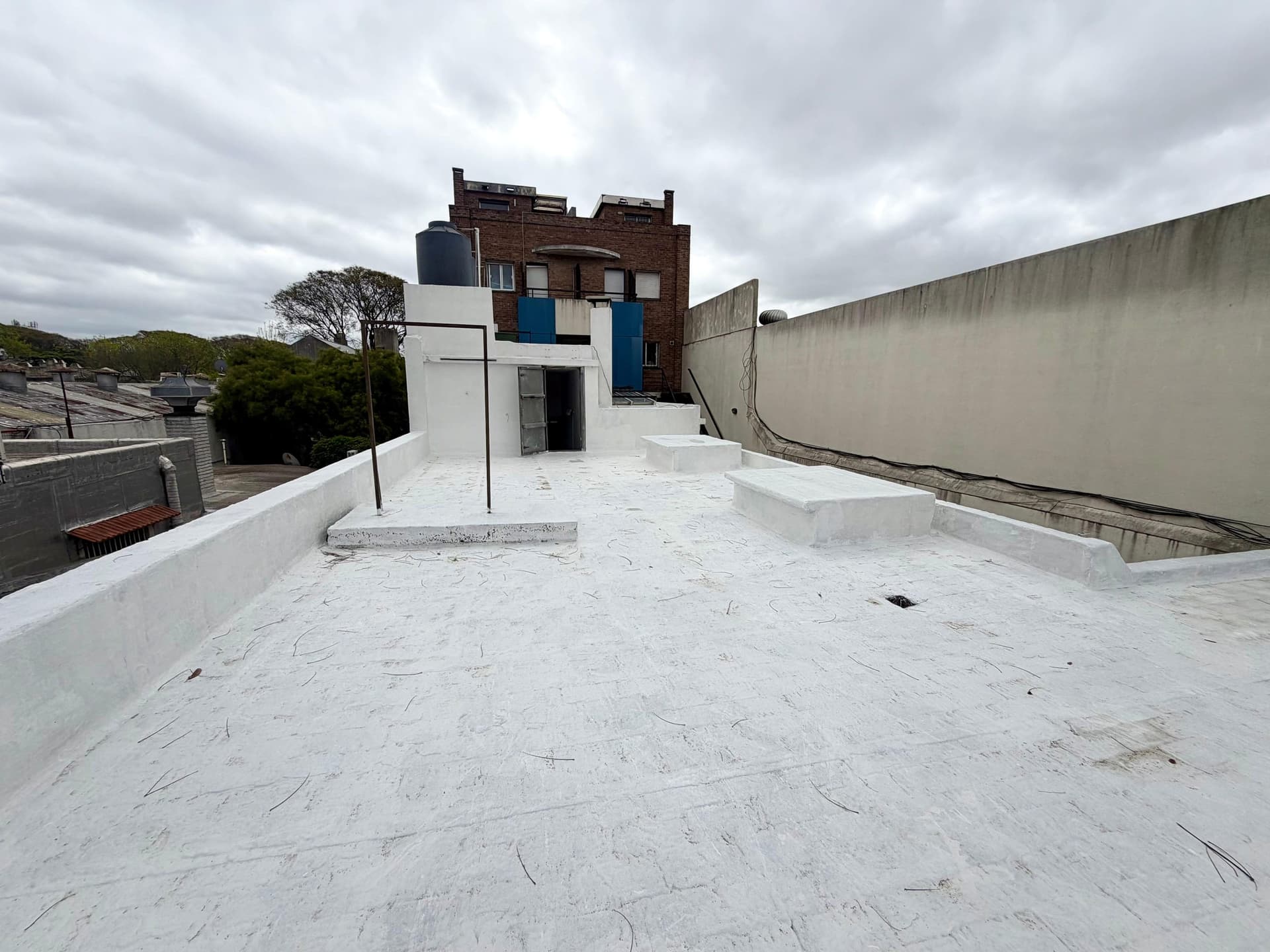 # Rooftop Terrace/Exterior Space
This is a spacious rooftop terrace featuring a newly waterproofed white surface with concrete finishes and raised edge detailing. The space adjoins a mixed-use building with red brick construction and blue accent panels, along with a modern white addition, offering potential for outdoor entertaining or development. The terrace provides open views of surrounding residential areas and is bordered by concrete perimeter walls and neighboring structures.