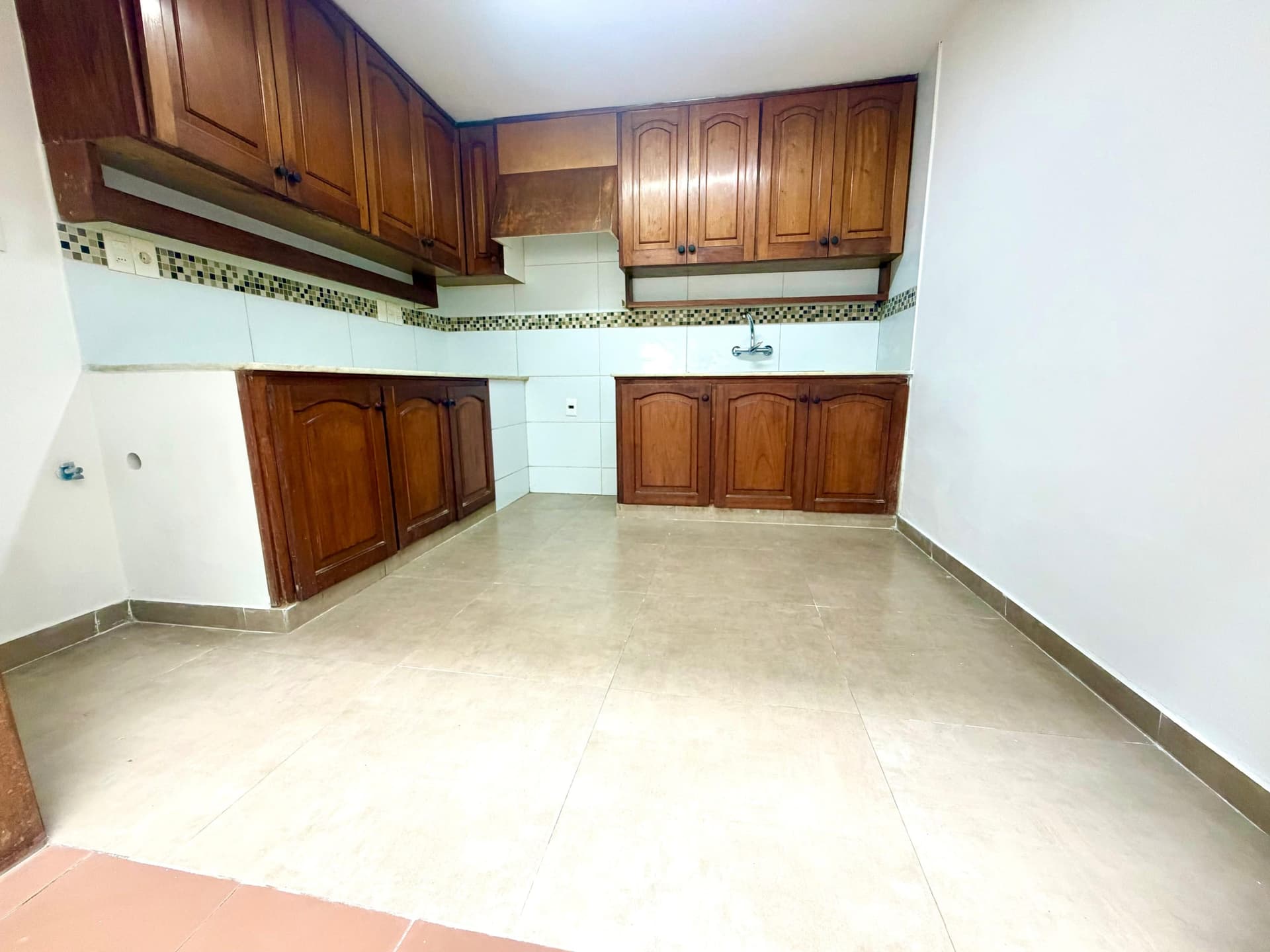 # Kitchen
This spacious kitchen features rich wood cabinetry with arched panel doors and decorative hardware throughout both upper and lower sections. The space includes white ceramic tile walls with a decorative mosaic tile border, a wall-mounted faucet, and neutral ceramic tile flooring, creating a clean and functional layout. The kitchen offers ample storage and counter space with good natural lighting from above.