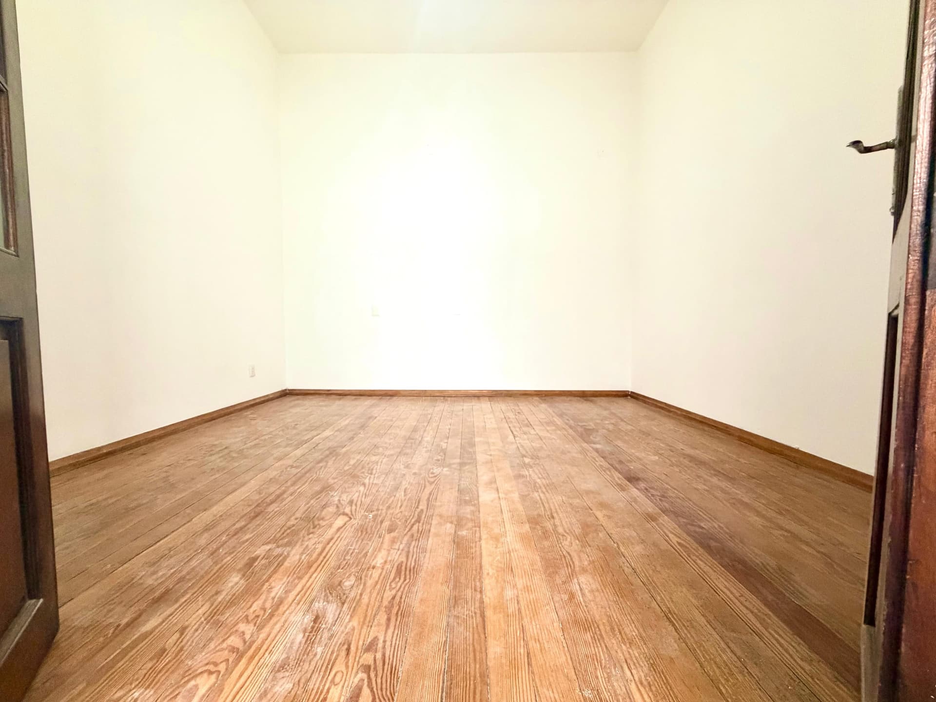 This is an empty bedroom or living space featuring hardwood flooring in a medium oak tone with a clean, uniform finish. The room has bright, neutral cream-colored walls, good natural lighting, and appears spacious with minimal obstructions. Two doorways with wood trim are visible on either side of the room, providing multiple entry points to the space.