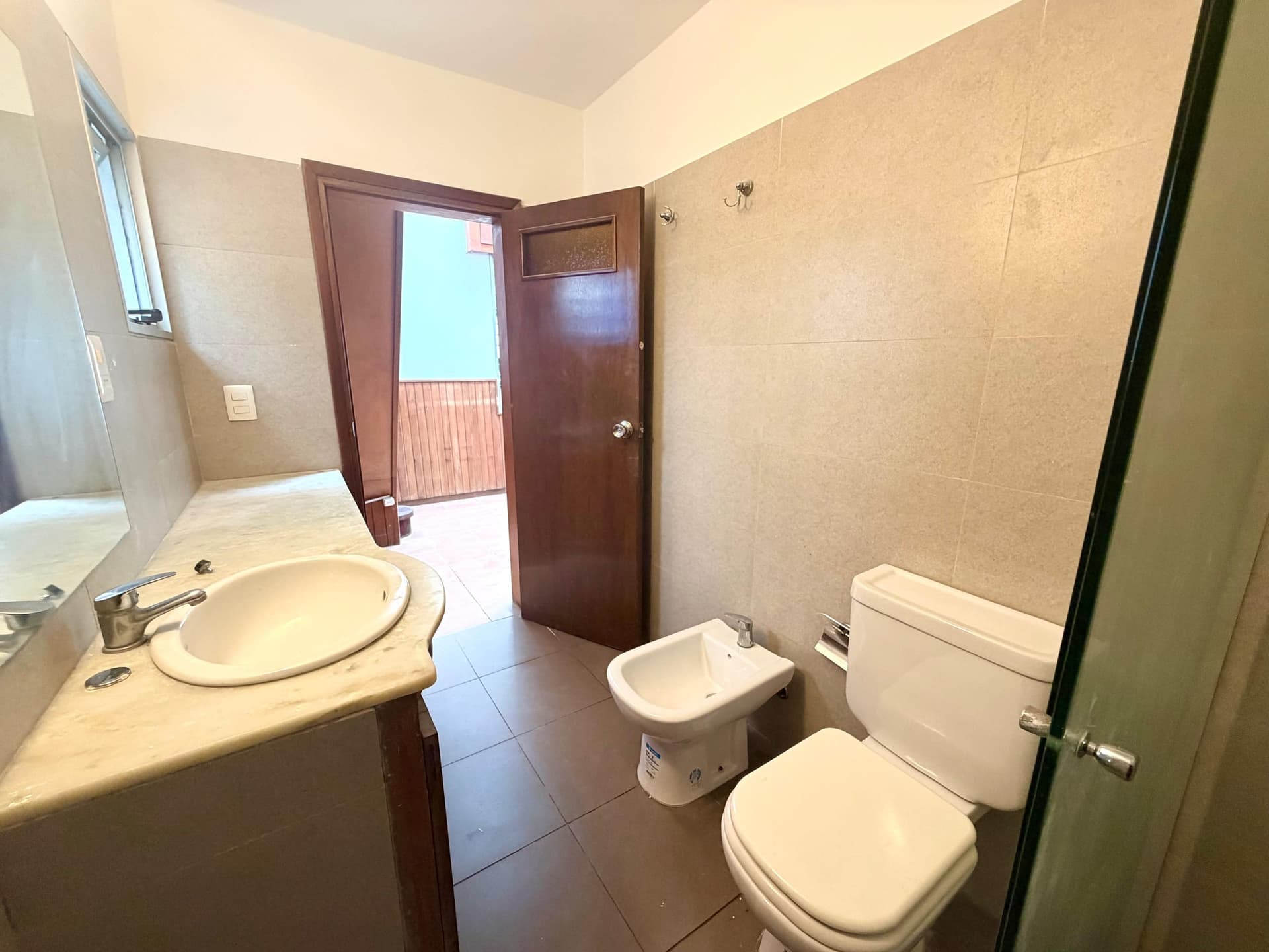 # Bathroom Description
This is a spacious bathroom featuring beige ceramic tile walls and dark tile flooring throughout. The room includes a round sink with vanity, a bidet, and a toilet, along with a small window providing natural light. A dark wooden door opens to an outdoor balcony/terrace area, and the bathroom is well-lit with bright ceiling fixtures, offering good functionality and ventilation.