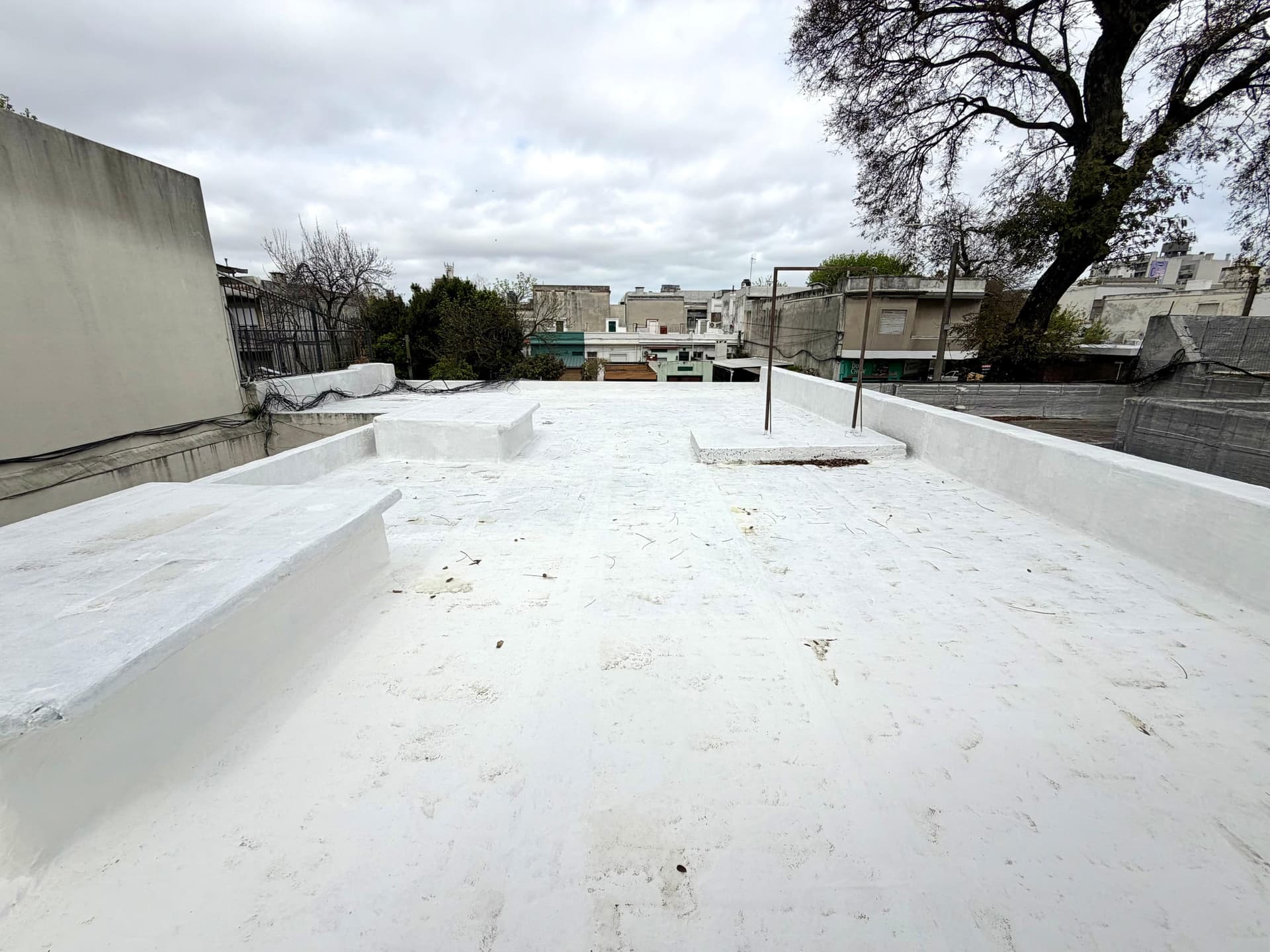 # Rooftop Terrace
This is a spacious rooftop terrace featuring a white waterproofed surface with concrete perimeter walls and metal railings for safety. The expansive outdoor space offers urban views overlooking neighboring Parisian buildings and mature trees, with ample room for development or outdoor living. The terrace appears to be in functional condition with good potential for renovation or customization.