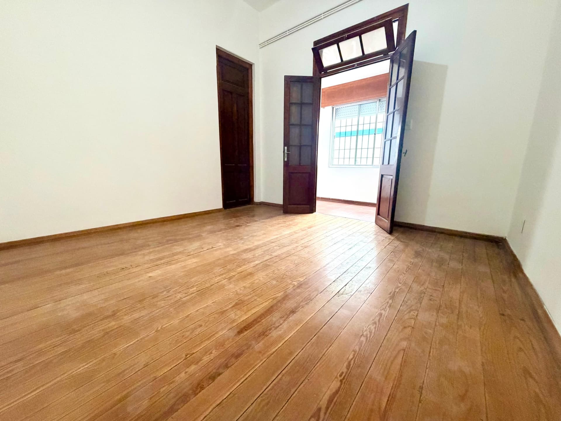 # Property Image Description
This is a spacious living room or bedroom featuring warm wood plank flooring in excellent condition throughout the space. The room is bright and airy with white walls, dark wood-framed doors, and a transom window above the main entrance that provides additional natural light. The open floor plan and clean, minimalist aesthetic make the space feel larger and move-in ready.