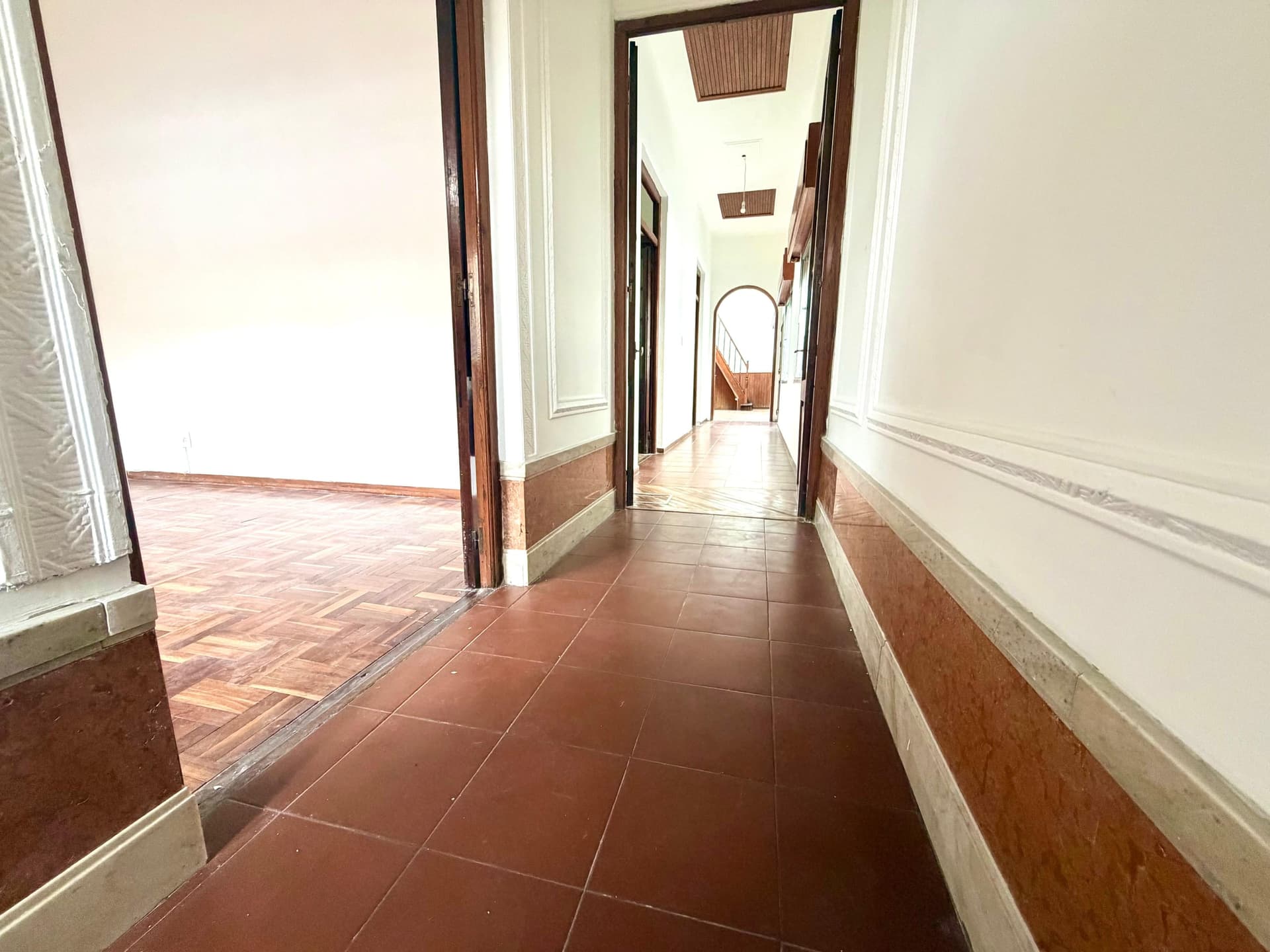 This is a hallway or corridor with terracotta tile flooring and cream-colored walls accented with a brown wainscoting base. The space features multiple wooden-framed doorways leading to adjacent rooms, with natural light streaming in from windows on the left side. The hallway appears to connect several areas of the property and shows good natural illumination, though the space would benefit from updating and maintenance.