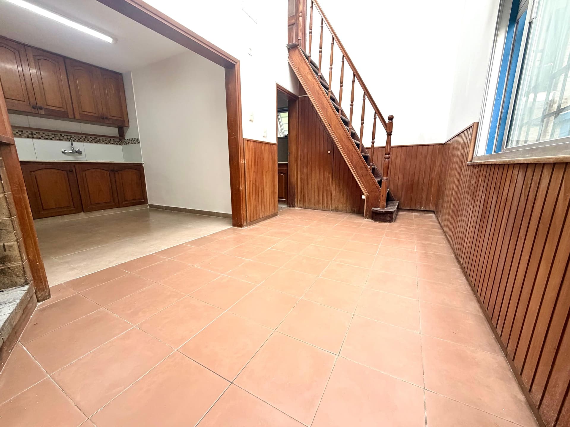 # Property Description
This is a spacious interior courtyard or patio area with terracotta tile flooring and distinctive wooden architectural elements throughout. The space features a wooden staircase with a carved handrail, wooden wall paneling, and wooden cabinetry/kitchen fixtures on the left side, creating a rustic, traditional aesthetic. The area is well-lit with natural light from windows on the right and artificial lighting overhead, offering good visibility and an open, airy feel despite the enclosed nature of the space.