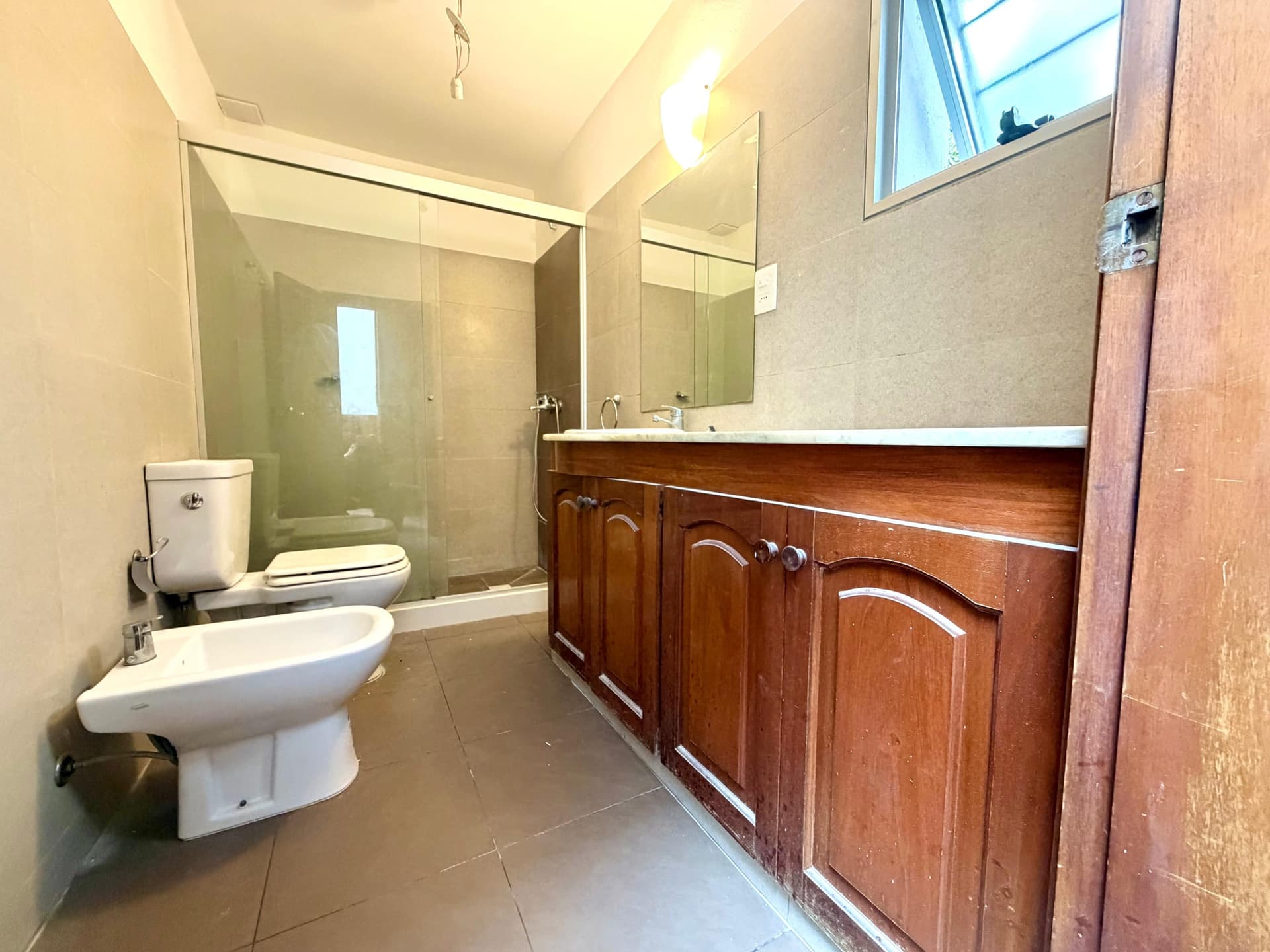 # Bathroom Description
This is a spacious master bathroom featuring a large walk-in shower with frameless glass enclosure and a separate soaking tub area. The room includes a wooden vanity cabinet with white countertop and dual mirrors, along with a white toilet and bidet fixture. The bathroom is finished with neutral tile flooring and walls, complemented by a high ceiling with recessed lighting and a window providing natural light. The wood cabinetry adds warmth to the contemporary design, and the layout offers excellent functionality and storage.