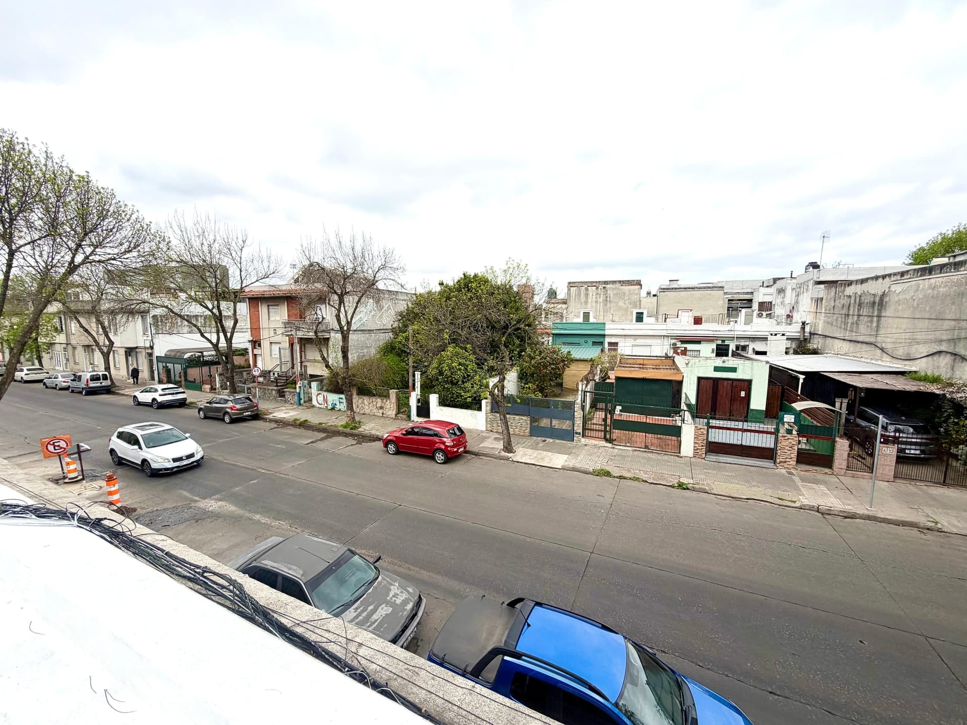 # Property Exterior - Street View
This image shows an elevated view of a residential neighborhood street with mixed architectural styles, including single and multi-family homes with varied facades in white, brick, and colored finishes. The street features on-street parking with several vehicles, mature trees throughout the area, and a mix of commercial and residential properties typical of an urban or suburban setting. The neighborhood appears to be in moderate condition with accessible street-level amenities and clear sightlines down the block.