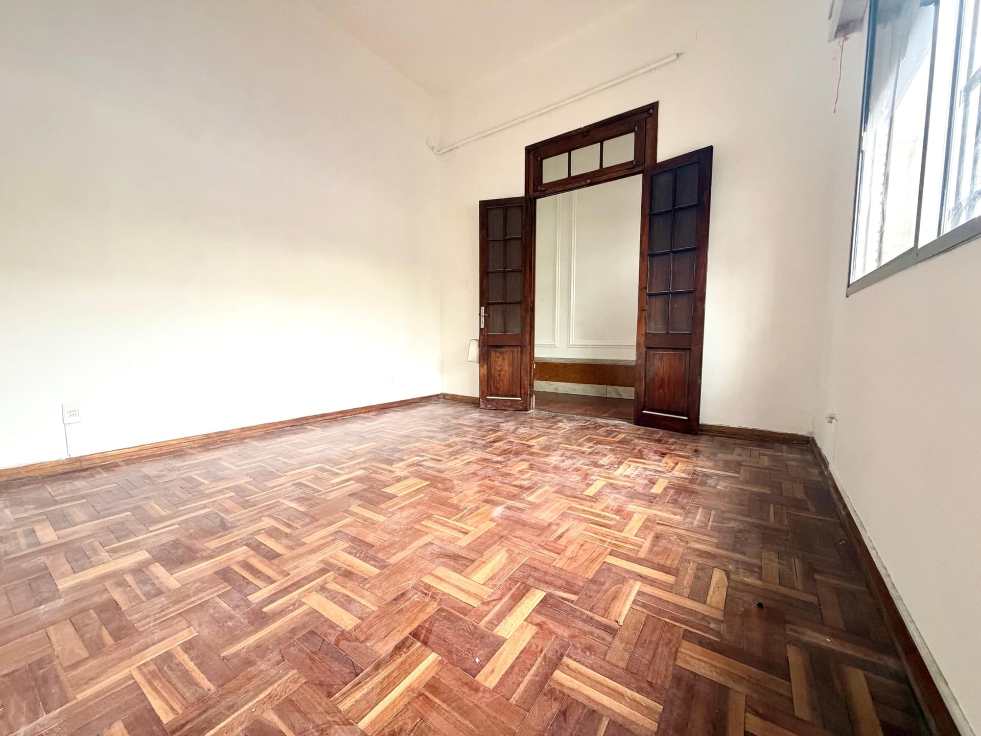# Property Description
This is a spacious bedroom or living room featuring beautiful herringbone parquet flooring in warm wood tones. The room is bright and airy with natural light from a large window on the right side, and it showcases distinctive dark wood-framed double doors with transom windows and decorative paneling, suggesting period or colonial-style architecture. The cream-colored walls and clean condition make this a versatile space with classic character and excellent natural lighting.