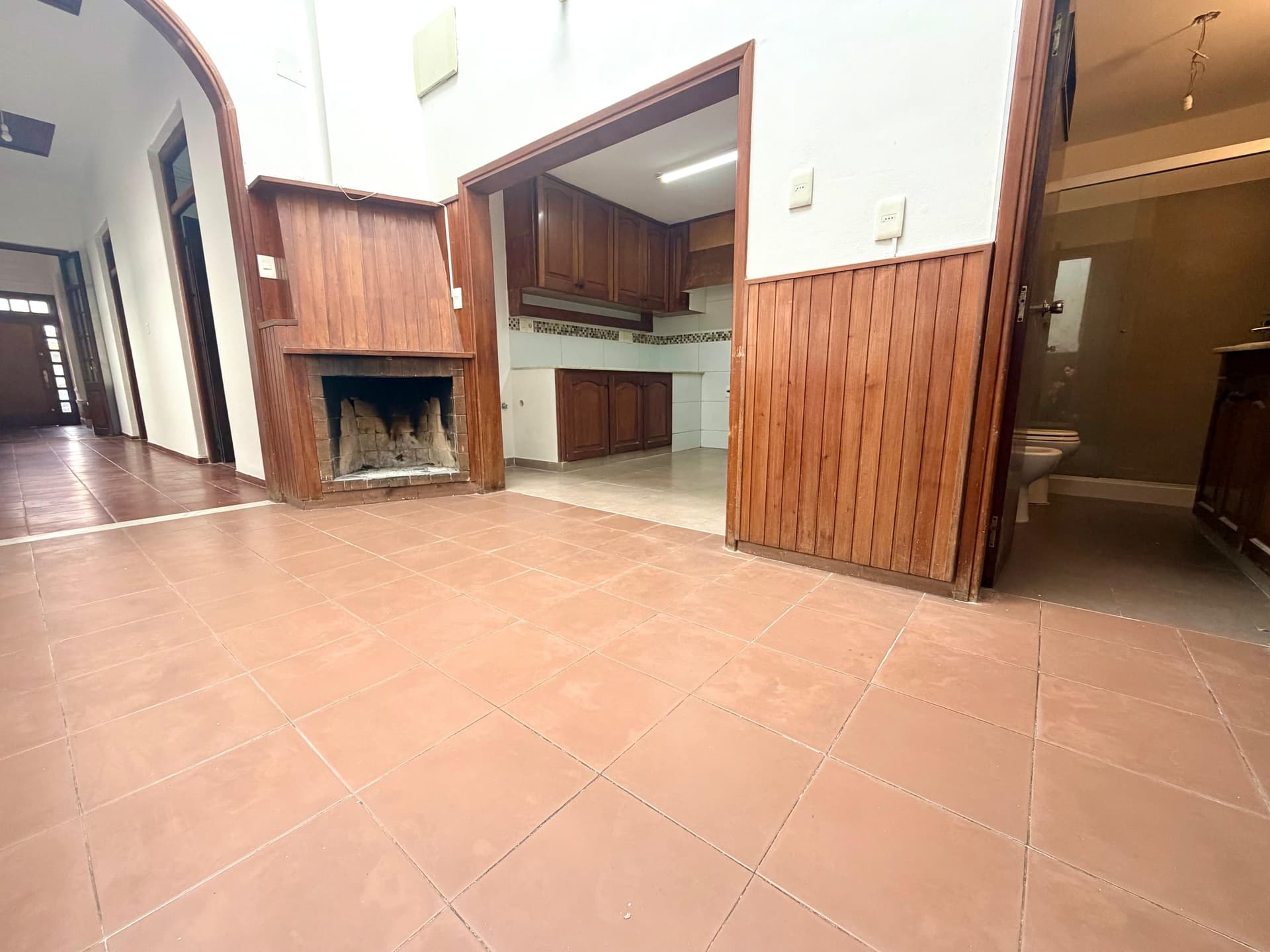 # Property Description
This is a spacious living/family room with terracotta tile flooring and high ceilings featuring white walls and warm wood accents. The space includes a decorative fireplace with wood paneling on the left side, wooden cabinetry and built-in features, and multiple wooden doors providing access to adjacent rooms and areas. The room is well-lit with natural light and appears to be in good condition, offering an open layout suitable for entertaining or family living.