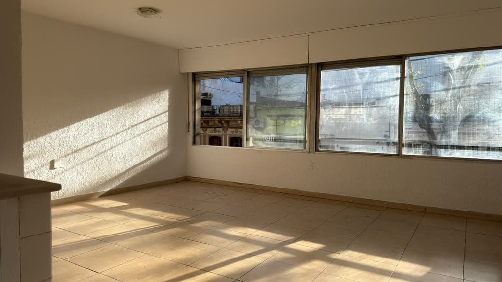 # Property Description
This is a bright, spacious living room or open-plan living area featuring light beige tile flooring and neutral cream-colored walls. The room is flooded with natural light from three large sliding glass windows that offer views of the surrounding neighborhood and outdoor spaces. The windows feature dark frames and appear to have security screens, while the clean, minimalist interior shows good condition with recessed ceiling lighting and a partial view of an adjacent room or kitchen area on the left side.