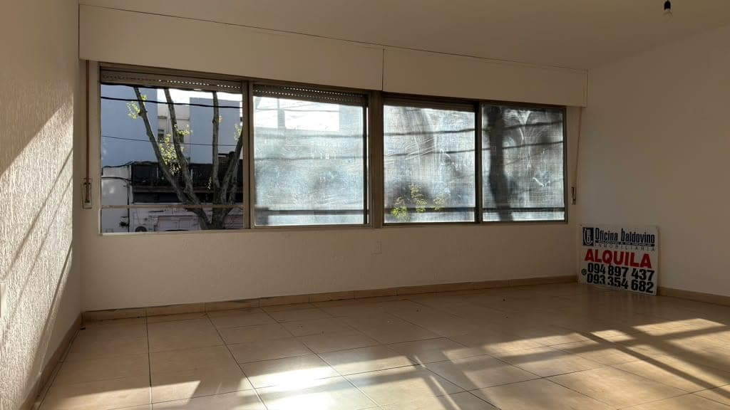 This is a bright, empty commercial or residential space featuring large multi-panel windows with brown frames that provide excellent natural light and views of the exterior. The room has neutral beige walls, light tile flooring, and clean, minimalist finishes suitable for office or retail use. An "ALQUILA" (for rent) sign is visible on the right wall, indicating this is a rental property.
WATERMARK: Oficina Deldoving